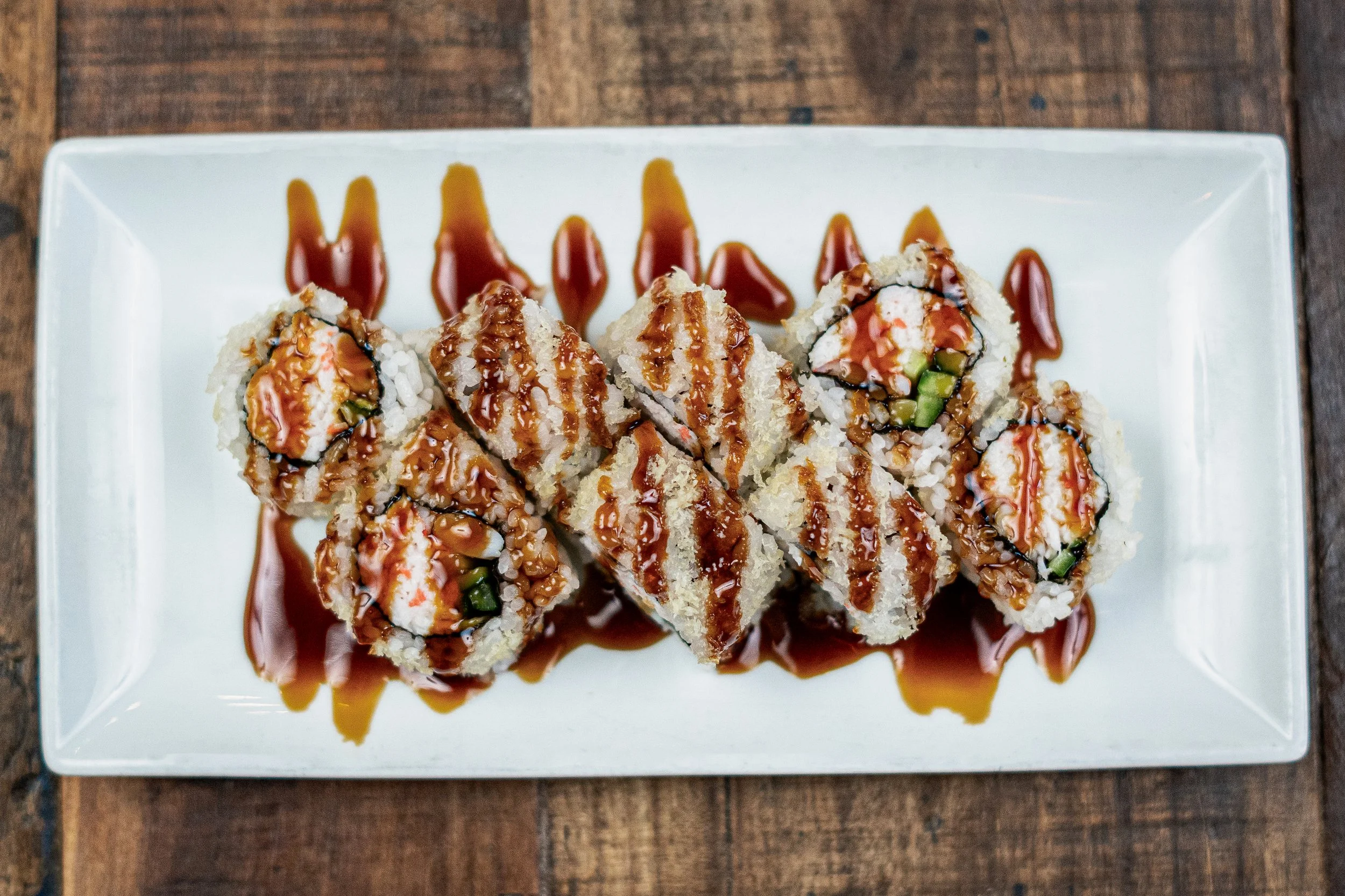 Sushi rolls drizzled with eel sauce on a rectangular white plate, garnished with rice and vegetables, on a wooden table.