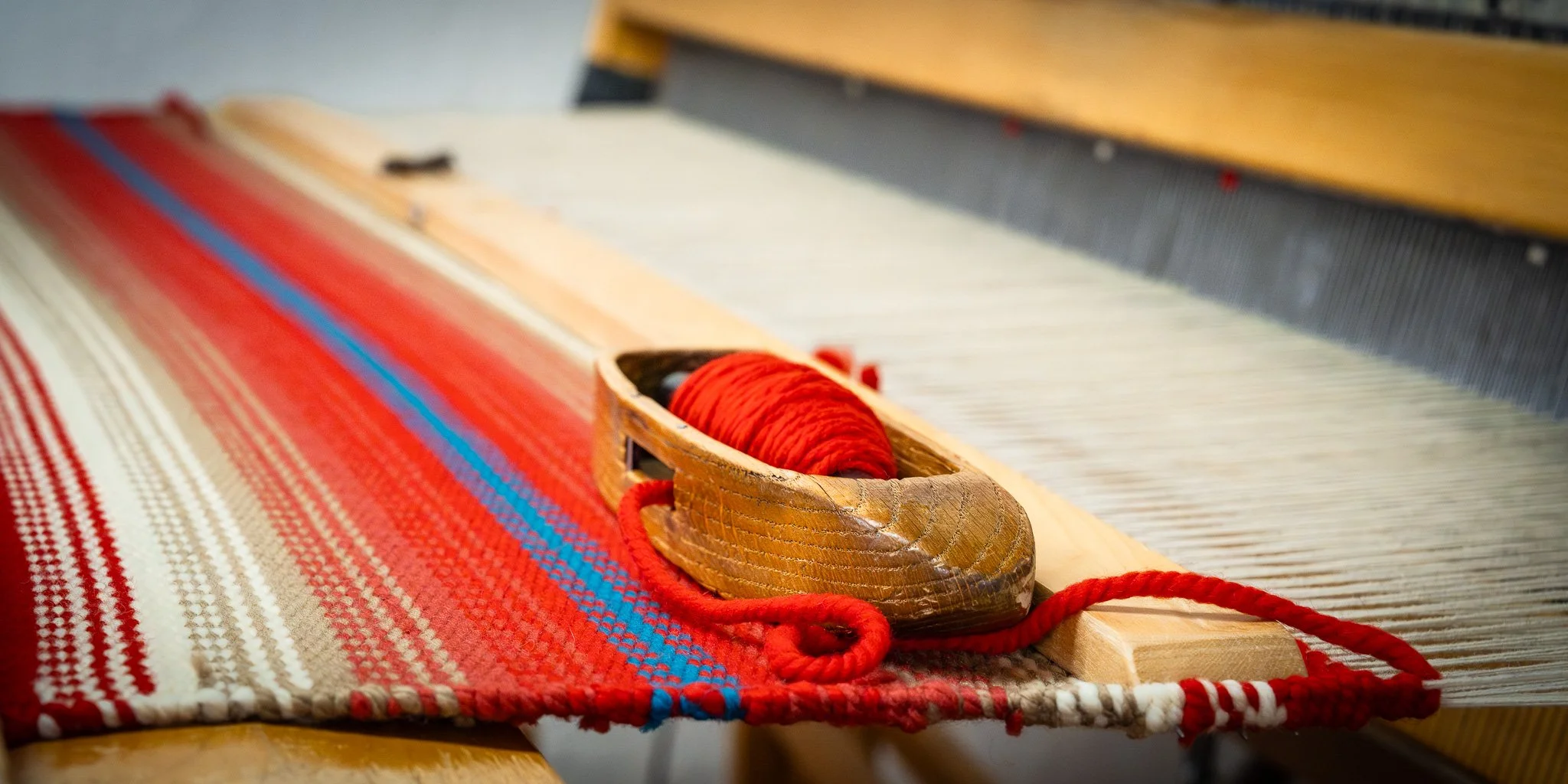 Close-up of a traditional handloom weaving machine with colorful woven fabric, showing the shuttle with red thread, and the wooden parts of the loom.