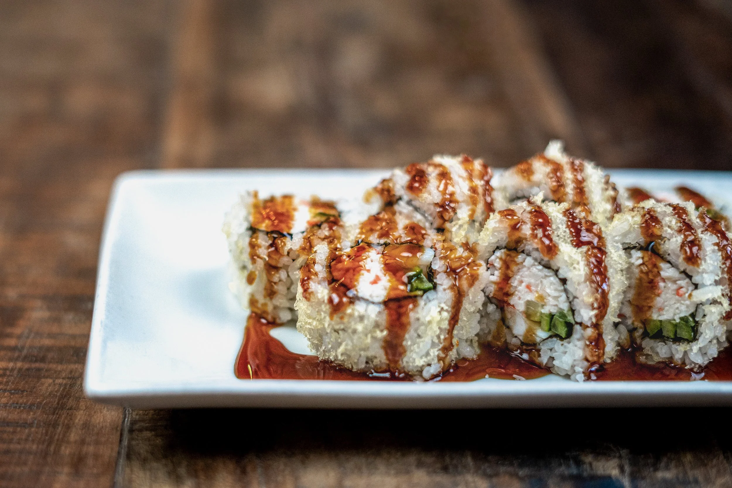Sushi rolls with white rice, fish, cucumber, and sauce, served on a white rectangular plate on a wooden surface.