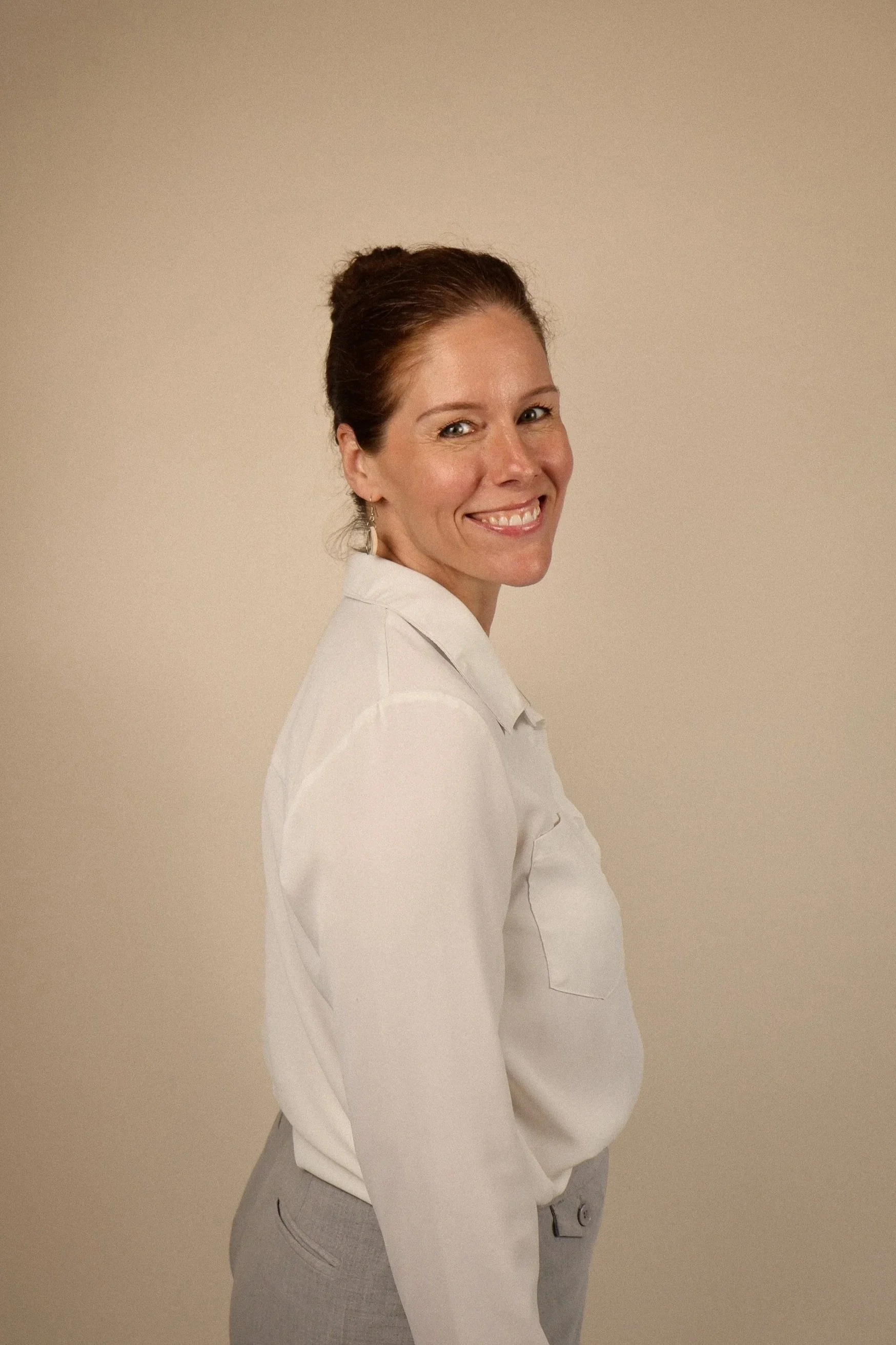 A woman with long, wavy brown hair smiling, wearing a light pink shirt and a white blazer, standing in front of a beige wall and a mirror.