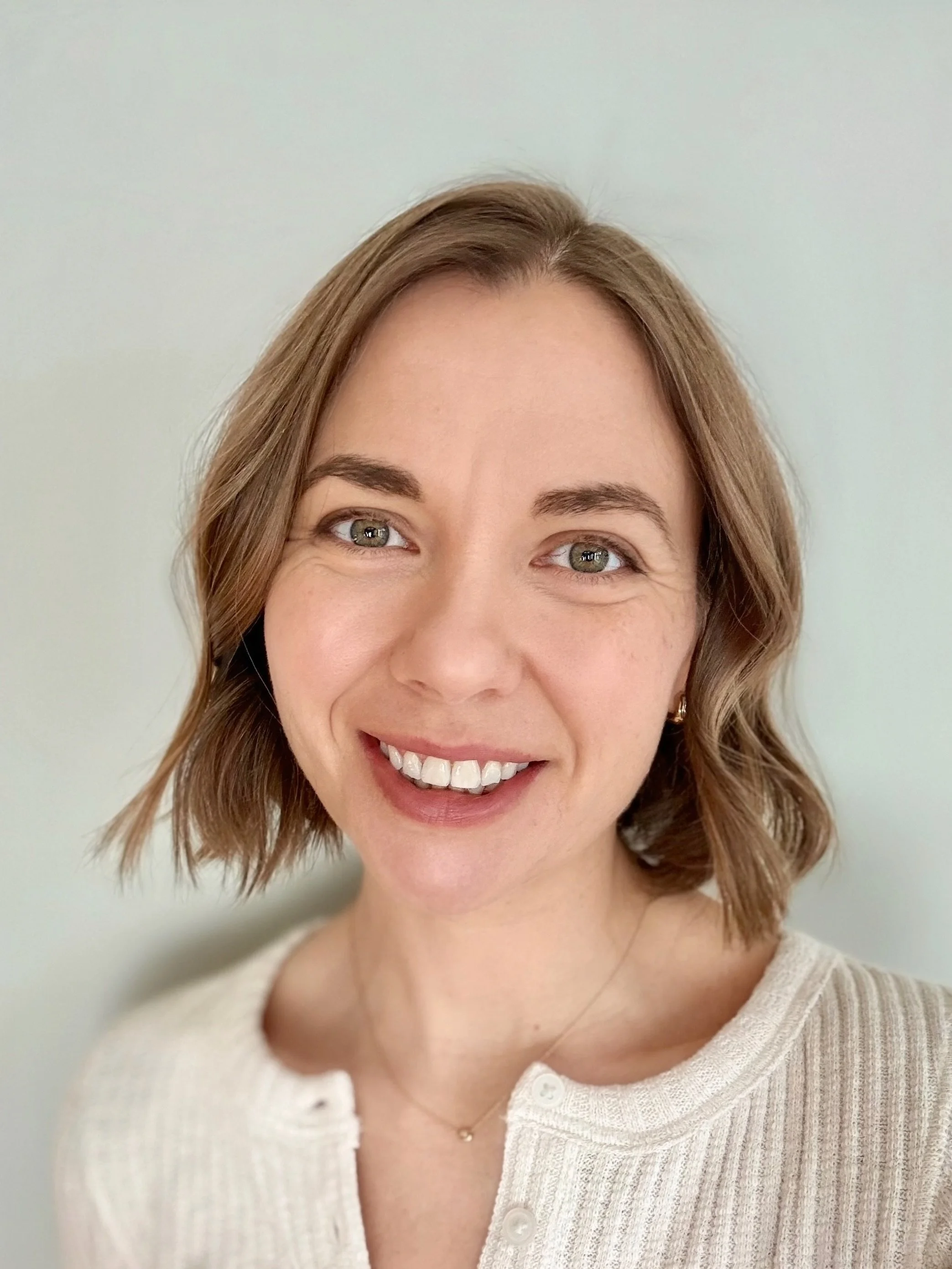 Close-up of a woman with shoulder-length light brown hair, smiling and looking at the camera, wearing a cream-colored top and a delicate necklace.
