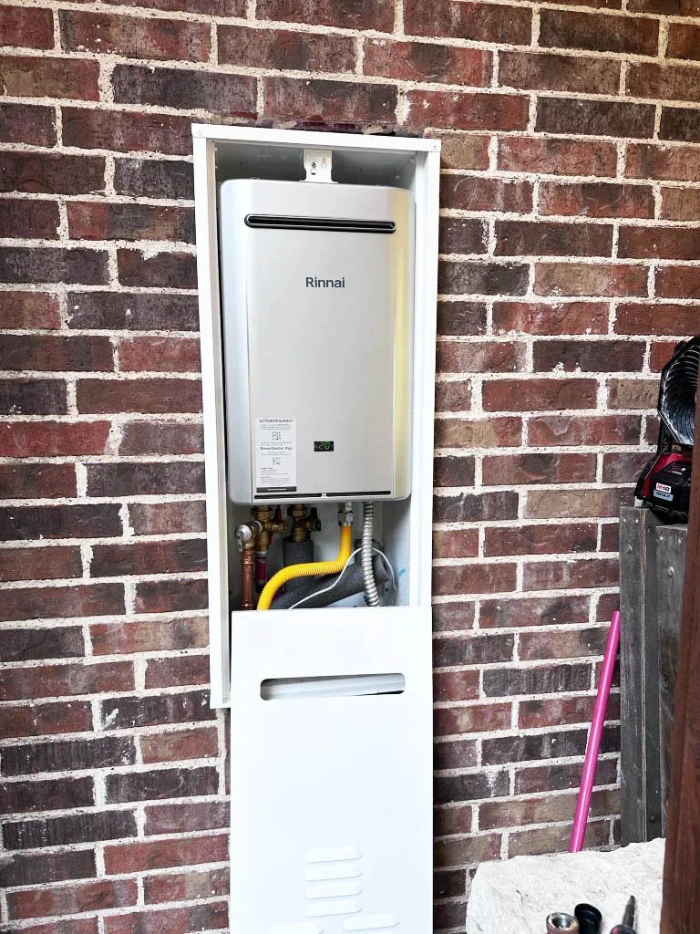 A Rinnai tankless water heater installed inside a white metal enclosure against a brick wall, with visible pipes and hoses.