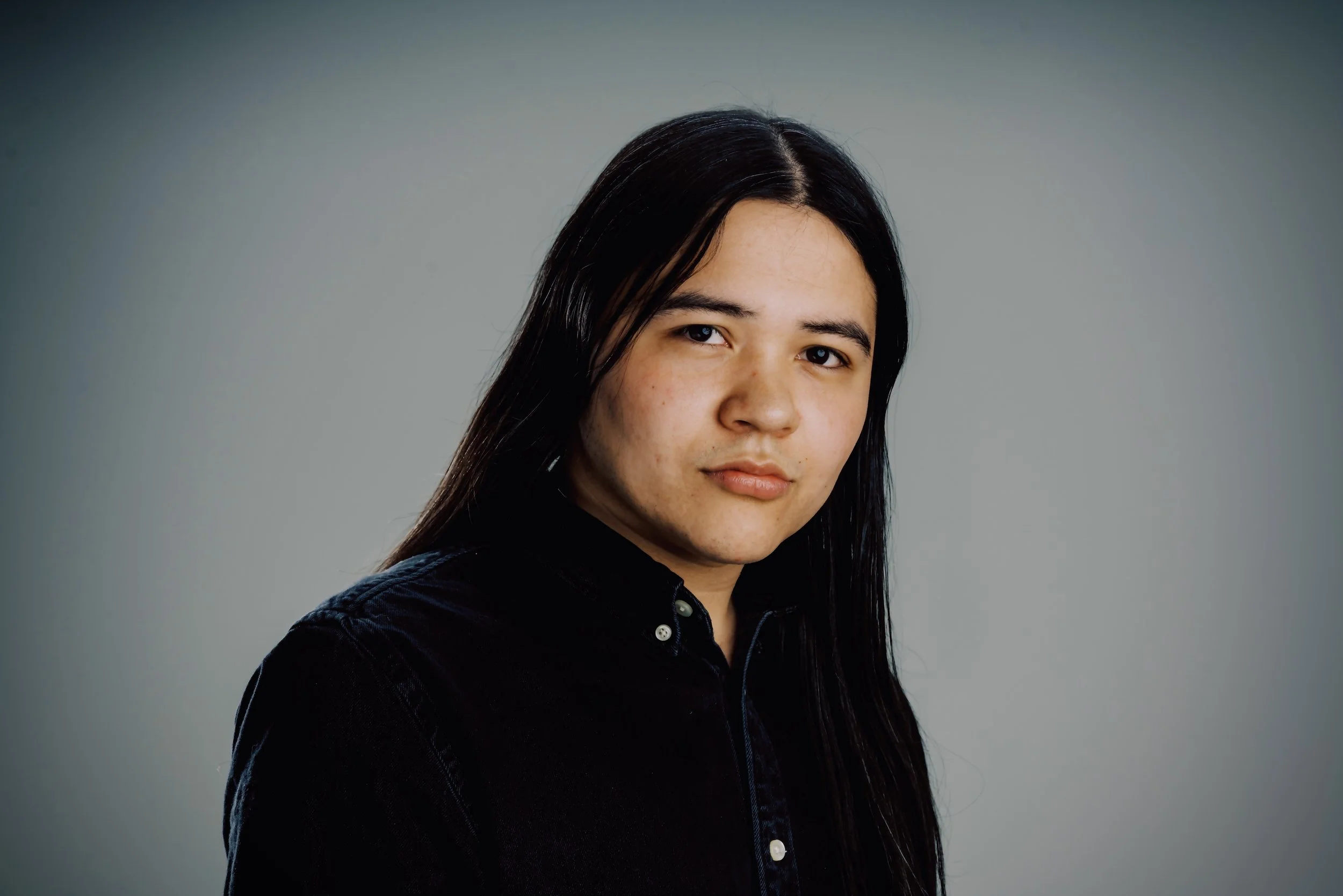 Portrait of a young woman with long dark hair, wearing a black shirt, against a plain gray background.