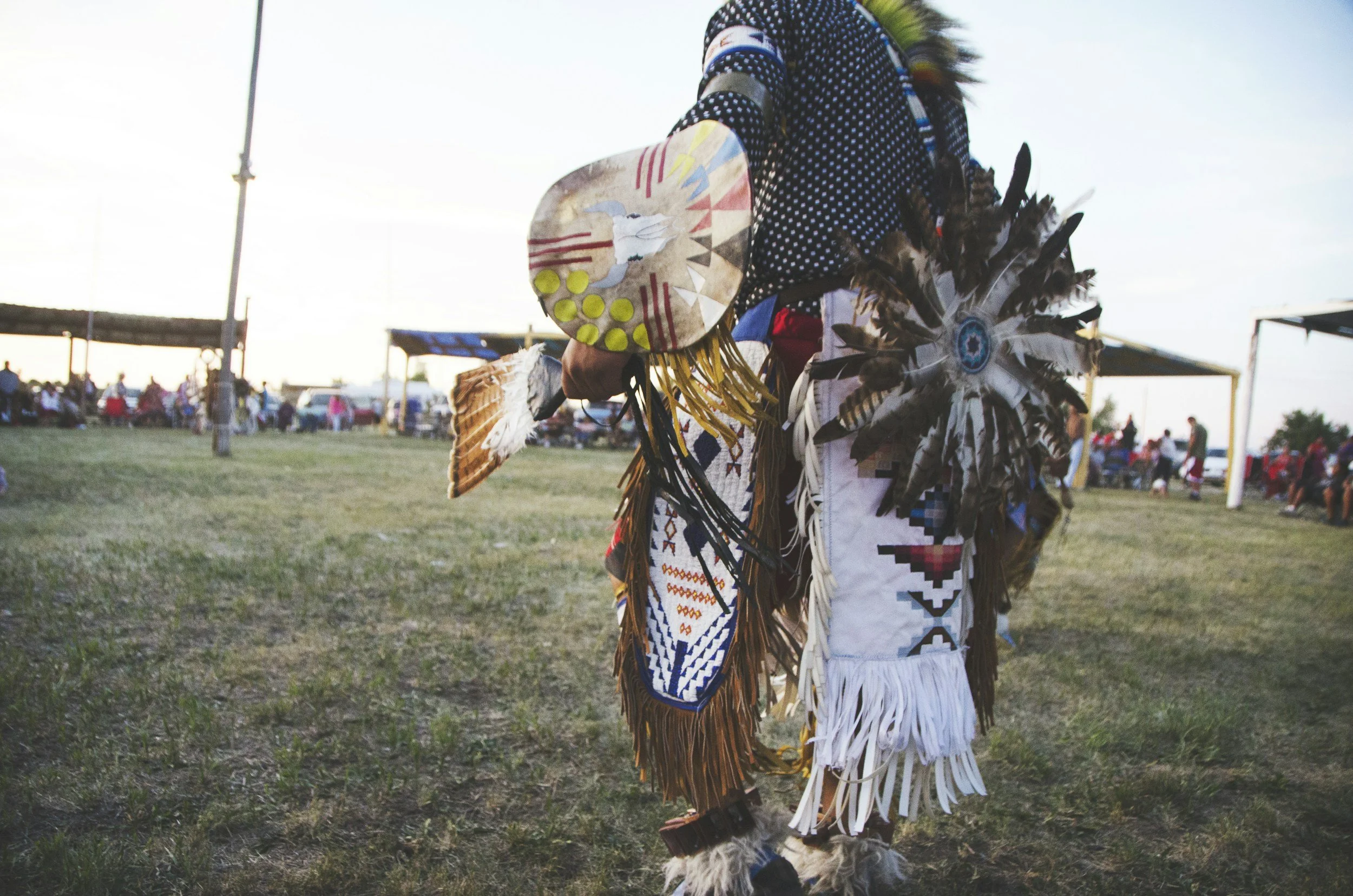 A person dressed in traditional Native American attire with feathered accessories at a cultural event outdoors, with a crowd in the background.