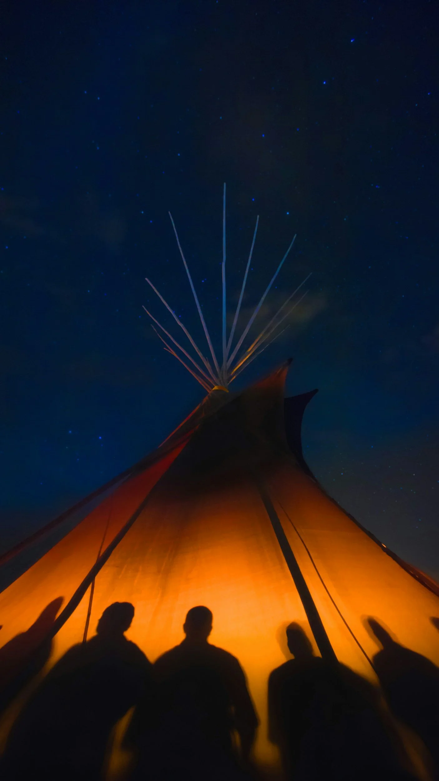 People sitting in a glowing orange tent at night, with a clear starry sky and fireworks in the distance.