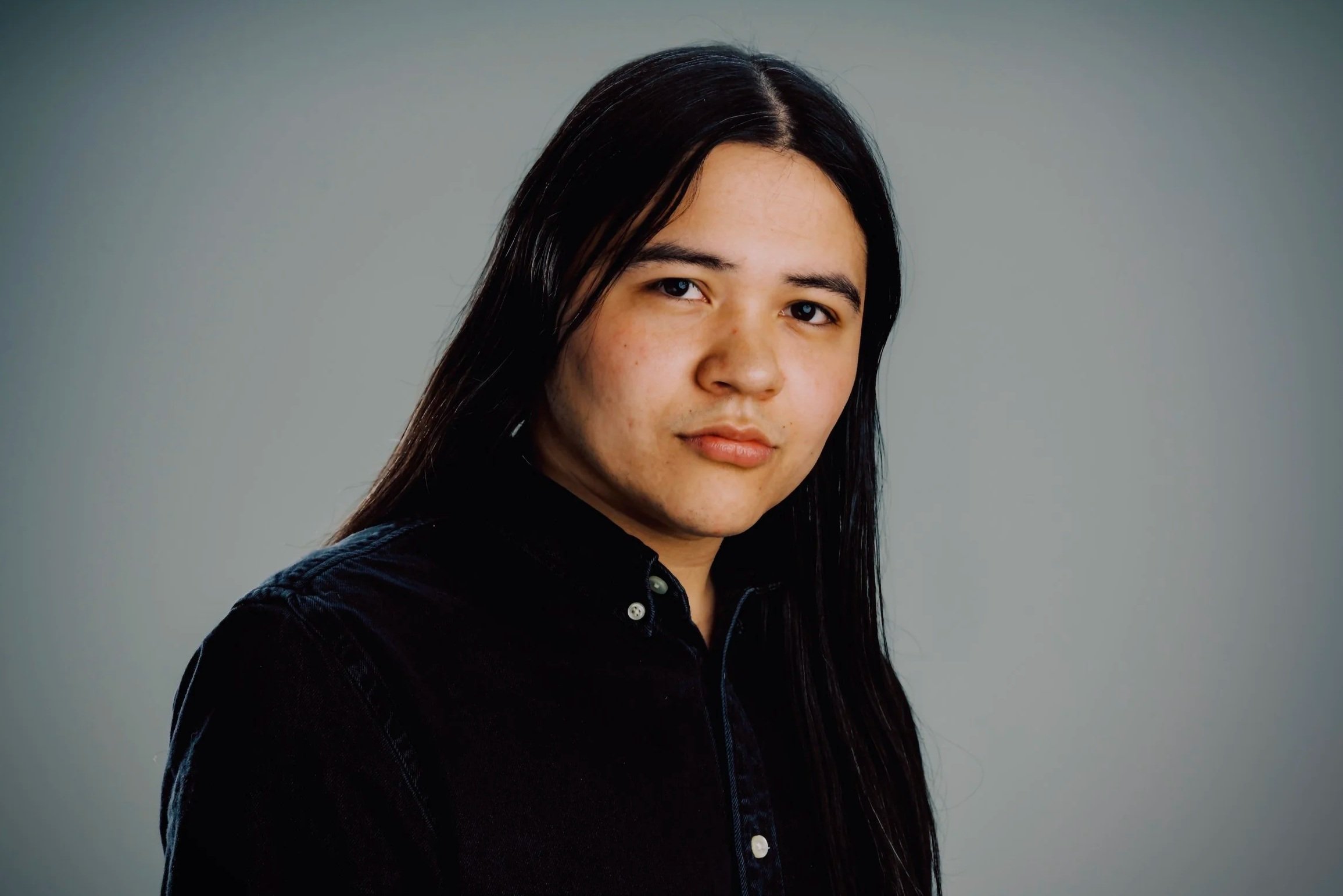 Portrait of a young woman with long dark hair, wearing a black shirt, against a plain gray background.