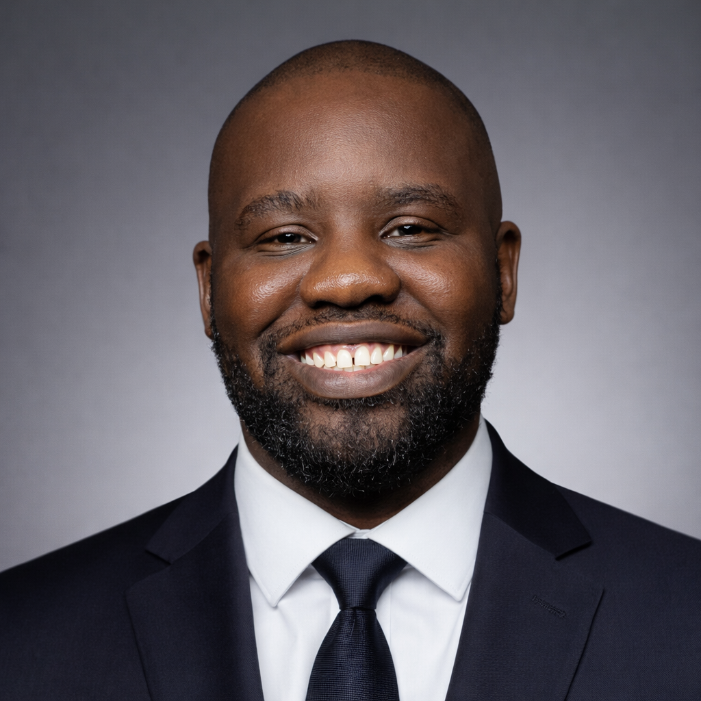 Headshot of a smiling man wearing a black suit, white shirt, and black tie against a grey background.
