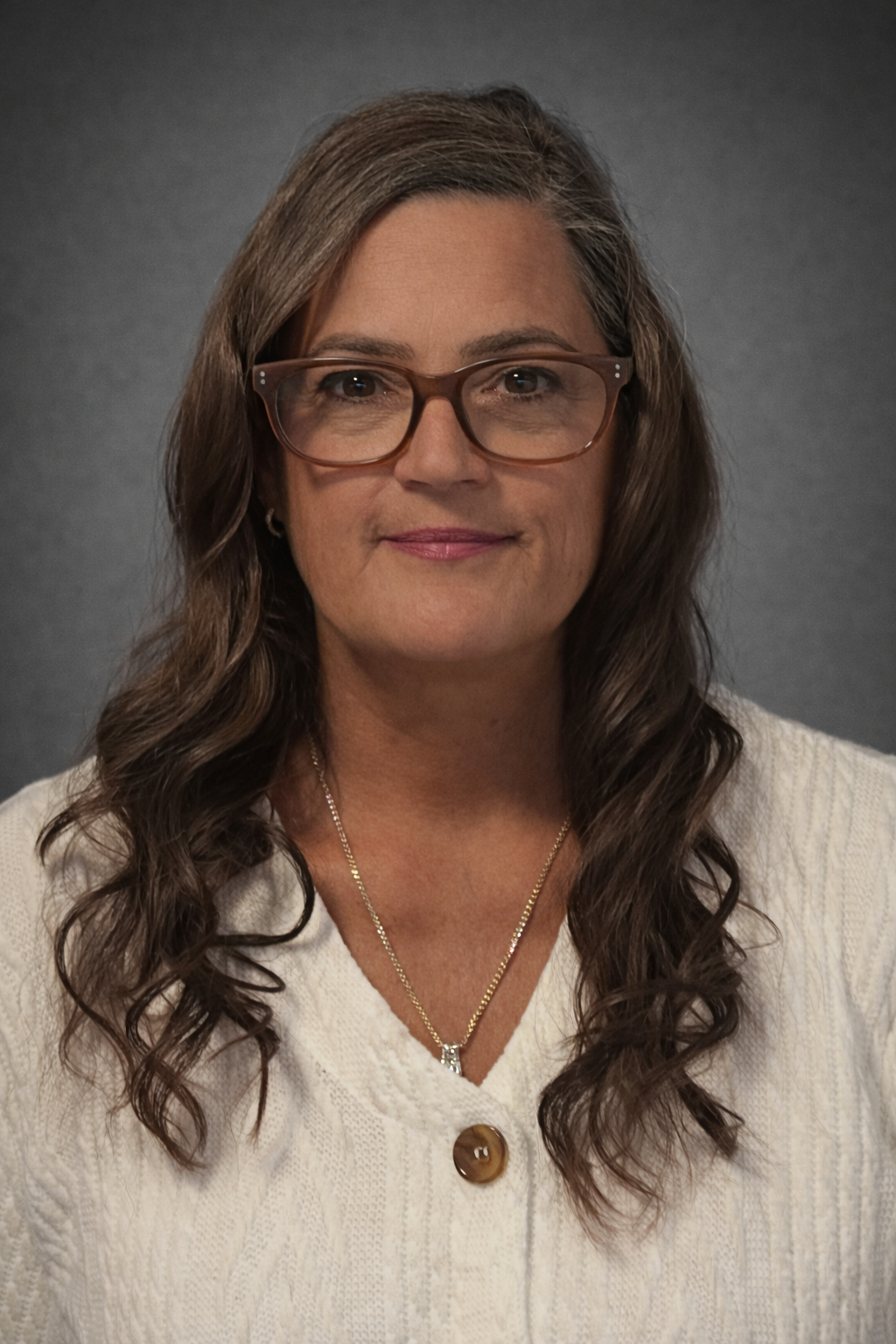 A middle-aged woman with long, wavy brown hair, wearing glasses, a white knitted sweater, a gold necklace, and earrings, smiling against a gray background.