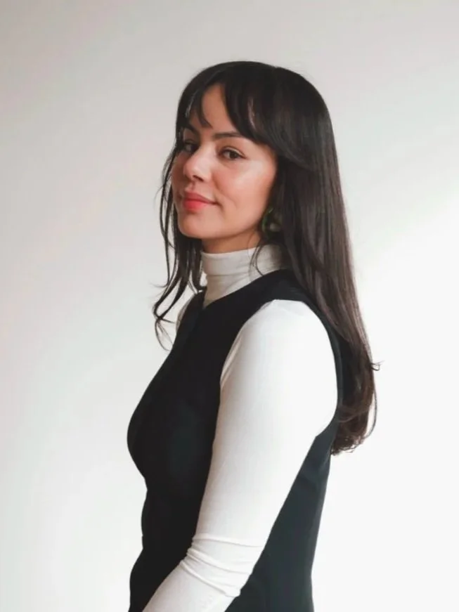 A woman with long dark hair wearing a white turtleneck and a black vest, standing against a plain white background.