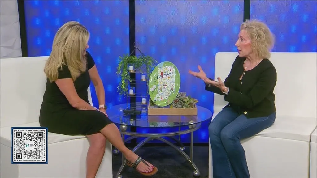 Two women having a conversation on a talk show set with a round glass coffee table in between. The woman on the left is sitting with her legs crossed, wearing a black dress. The woman on the right is gesturing with her hands, wearing a black top and blue jeans. There are decorative items on the table, including a round green sign and a small plant, with a blue illuminated background.