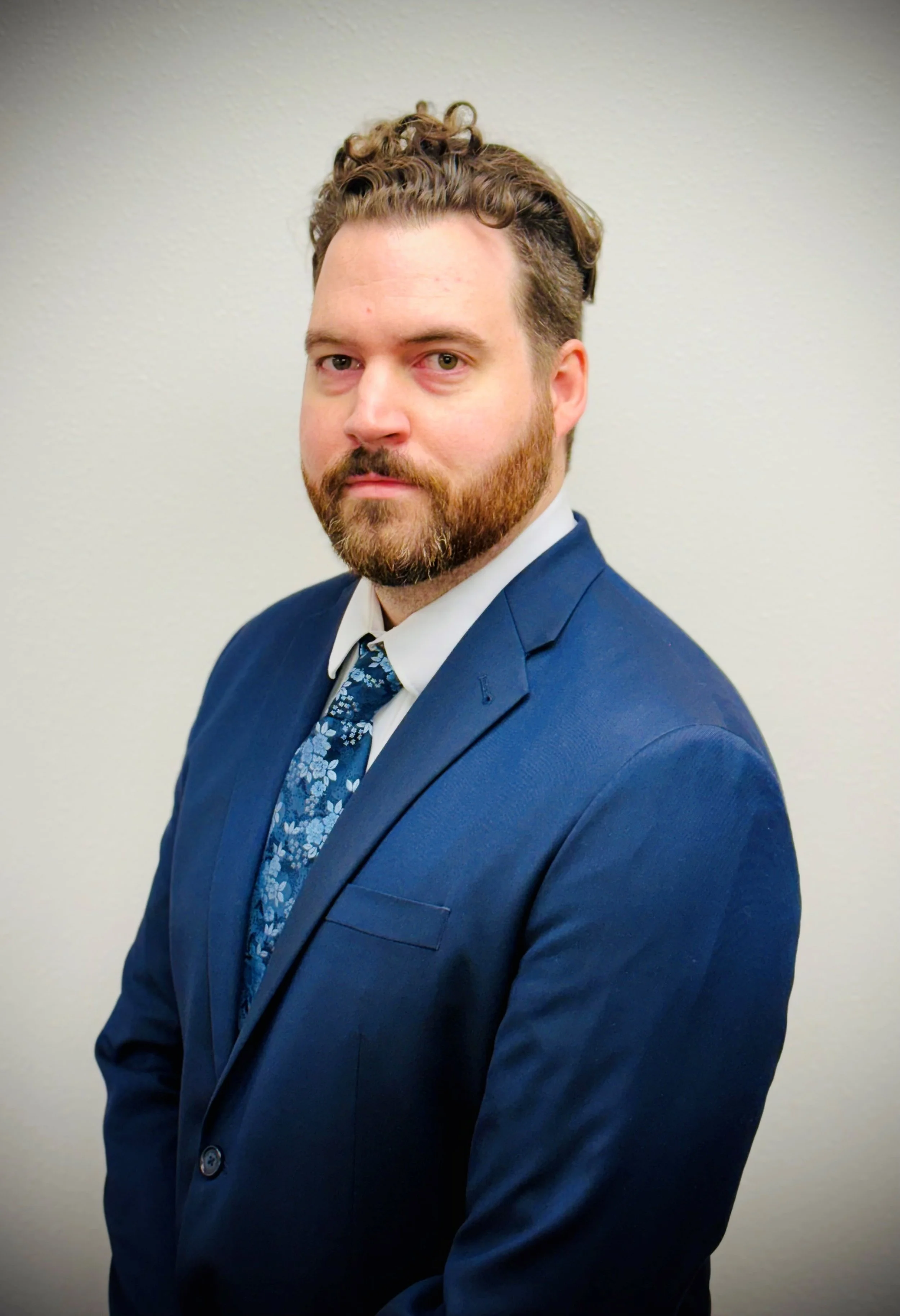 Man in a blue suit and floral tie against a plain background.