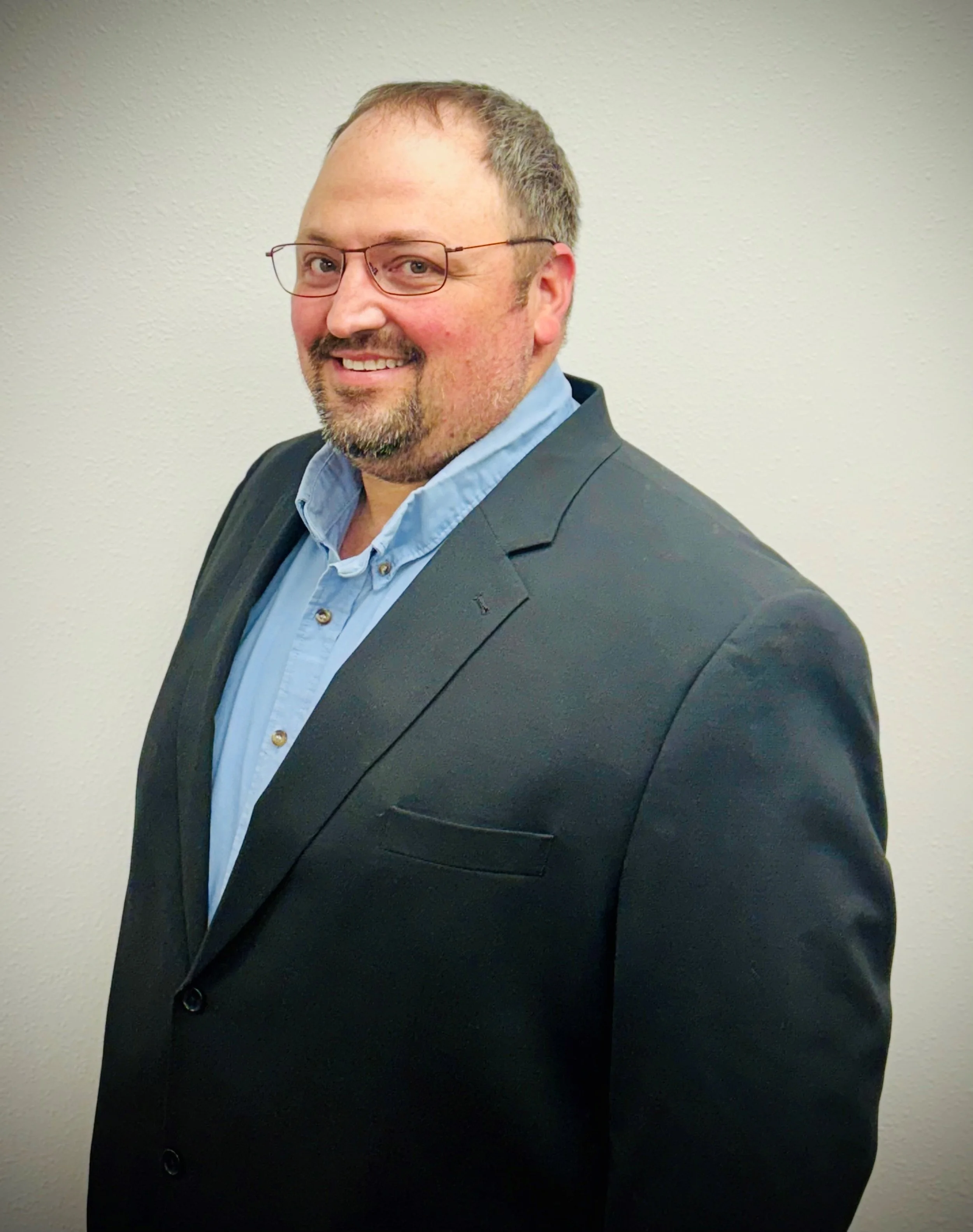 Man in a black suit and blue shirt smiling, standing against a white background.