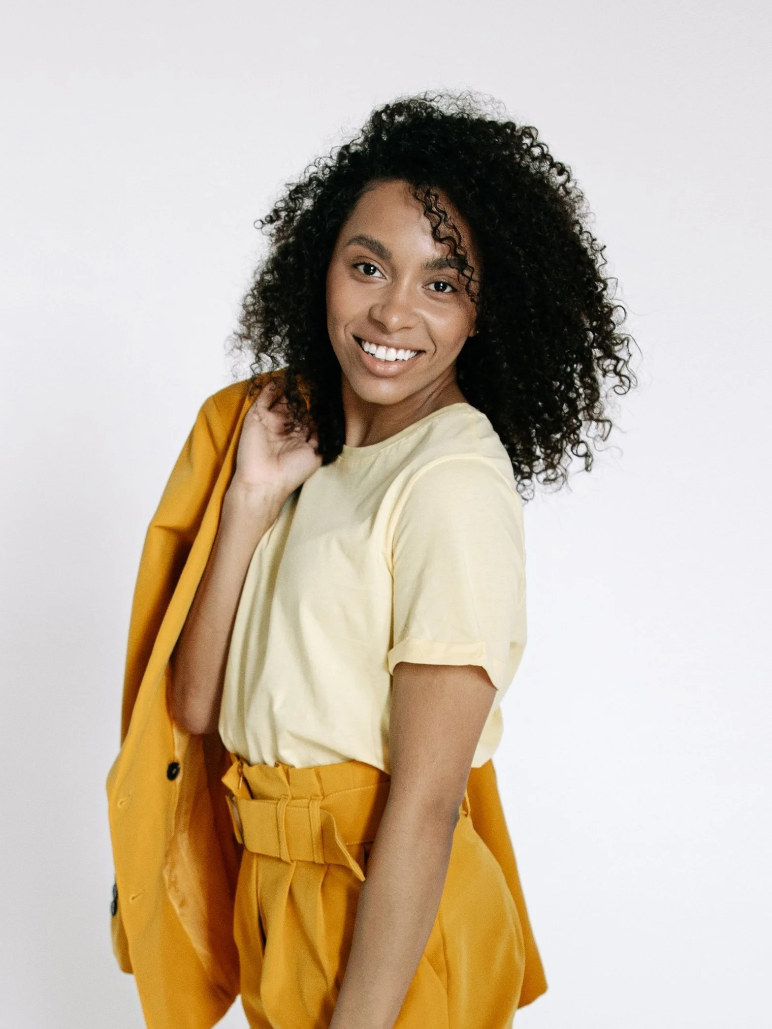 Smiling woman with curly hair holding an orange jacket draped over her shoulder, wearing a matching high-waisted skirt and a light-colored shirt.