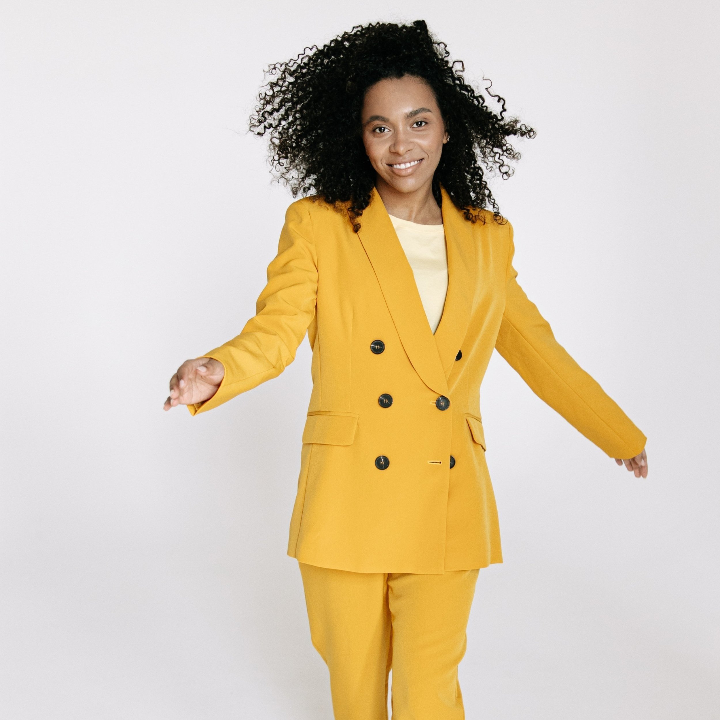 Person wearing a yellow suit with curly hair, posing with arms outstretched against a white background.
