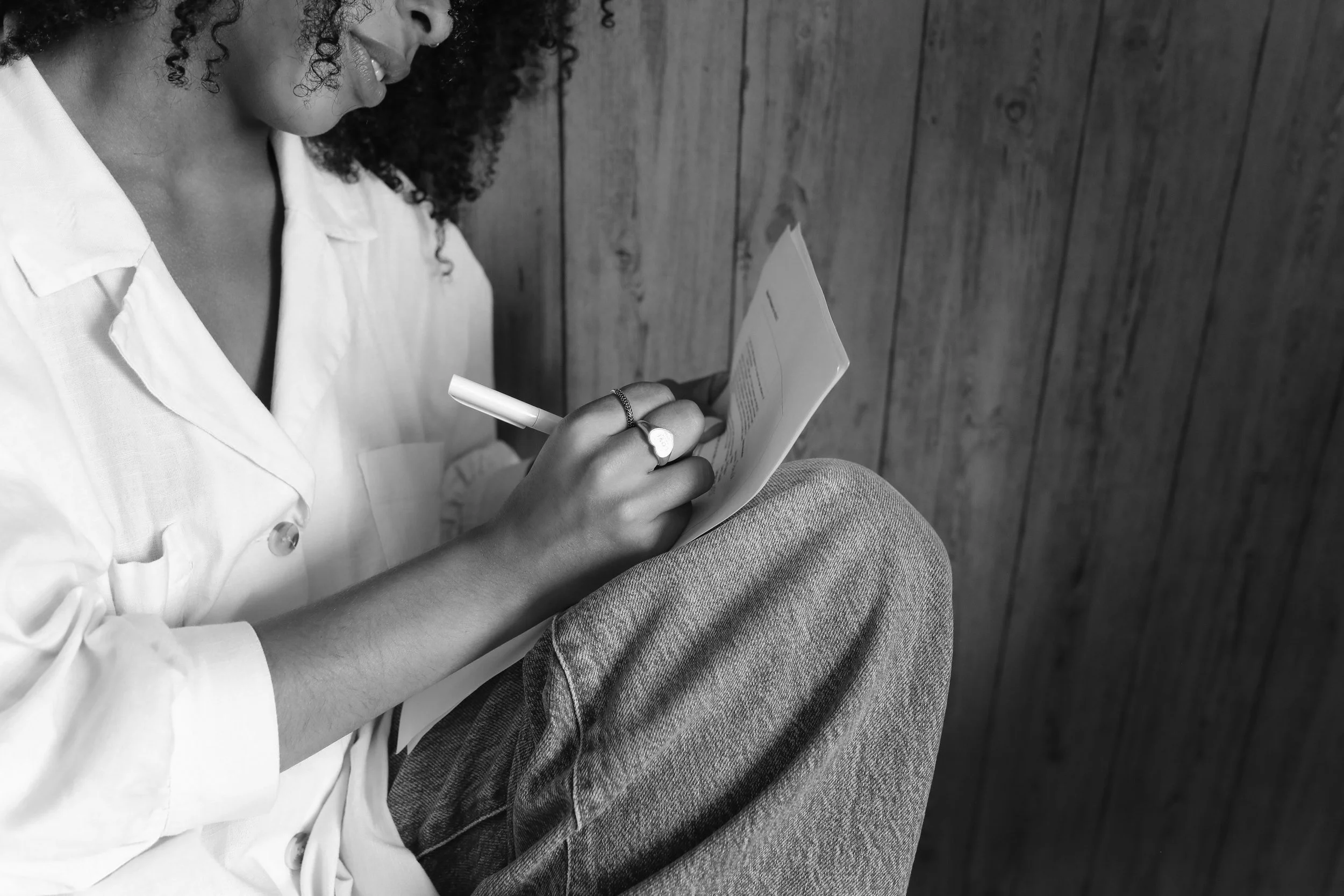 Black and white image of a woman in a white shirt and jeans writing on a notepad with a pen, sitting against a wooden wall.