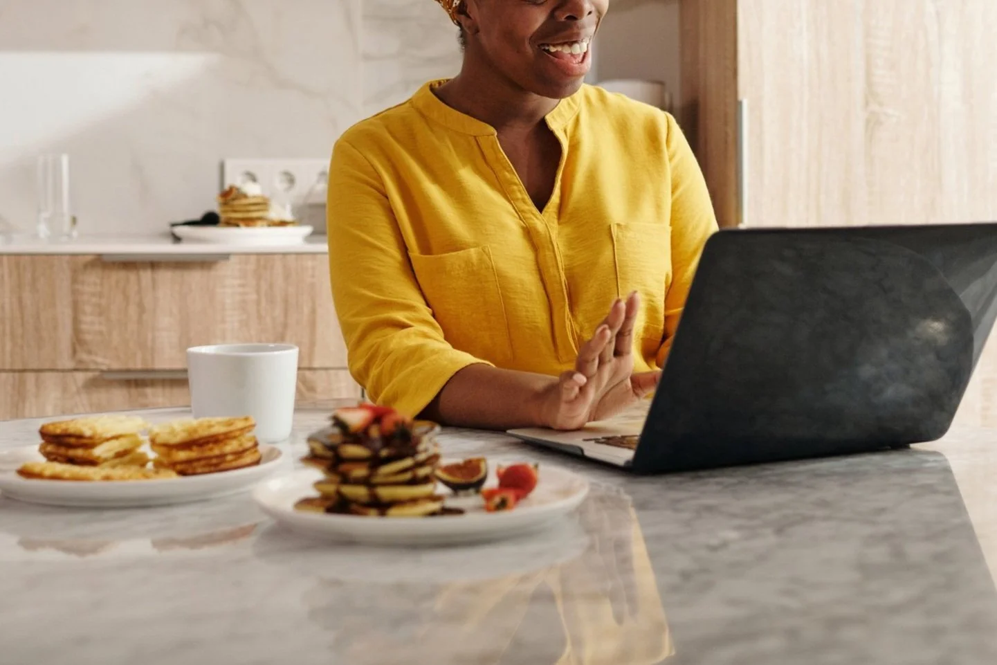 Person in a yellow shirt sitting at a kitchen counter working on a laptop with plates of pancakes and a cup on the side.