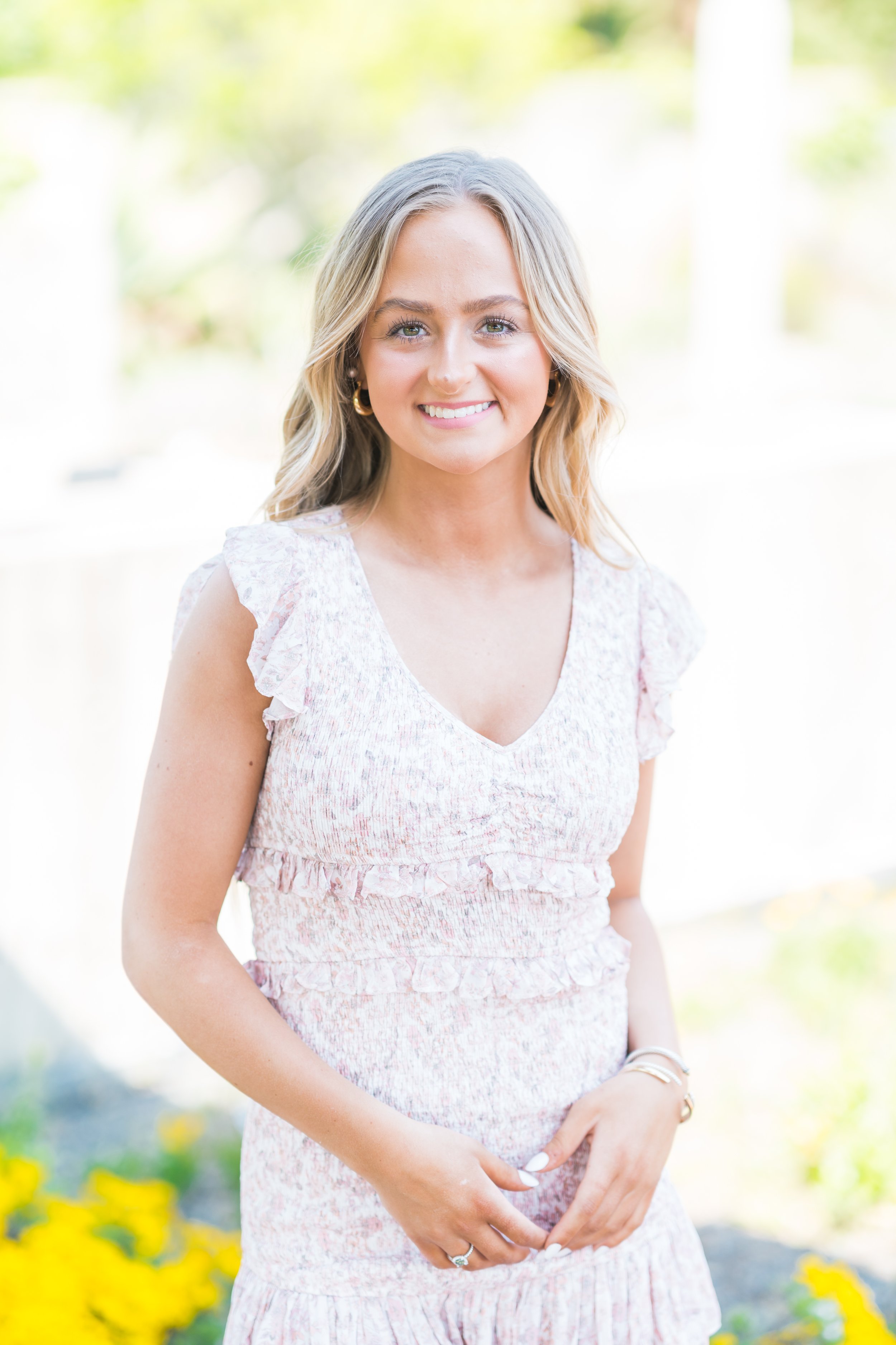 Smiling woman in a light floral dress standing outdoors with blurred greenery in the background.