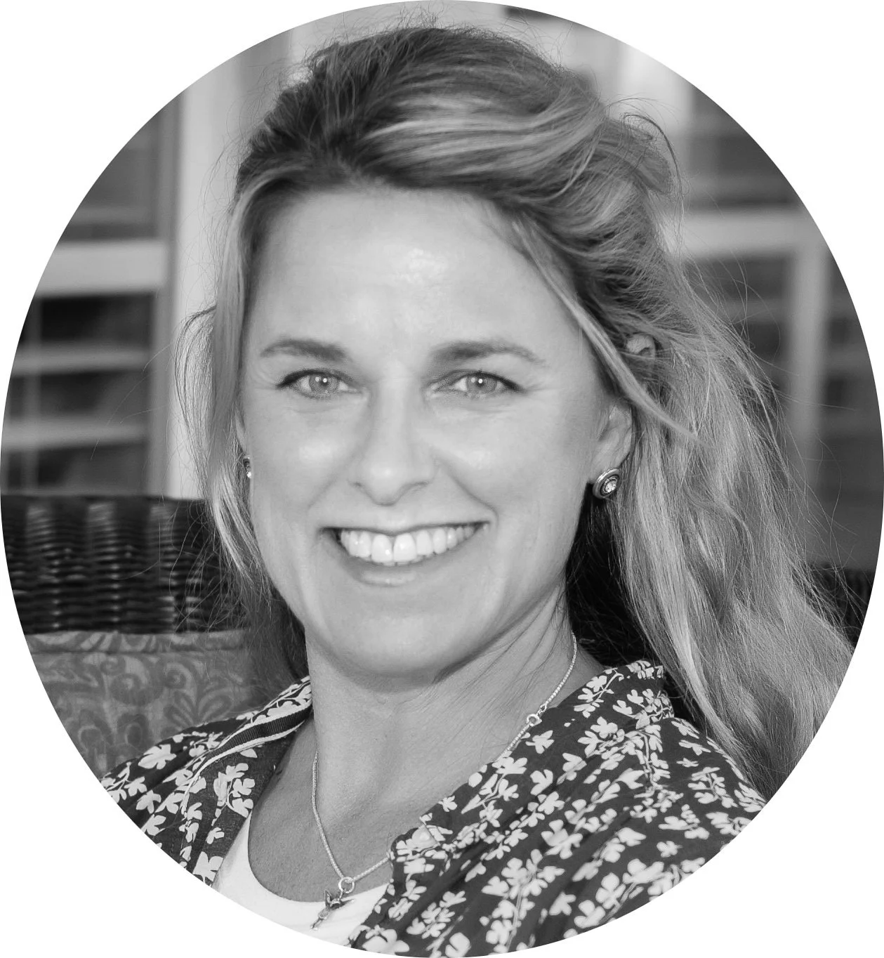 Black and white portrait of a smiling woman with wavy light-colored hair, earrings, and a floral patterned jacket, seated on a wicker chair with a blurred background.