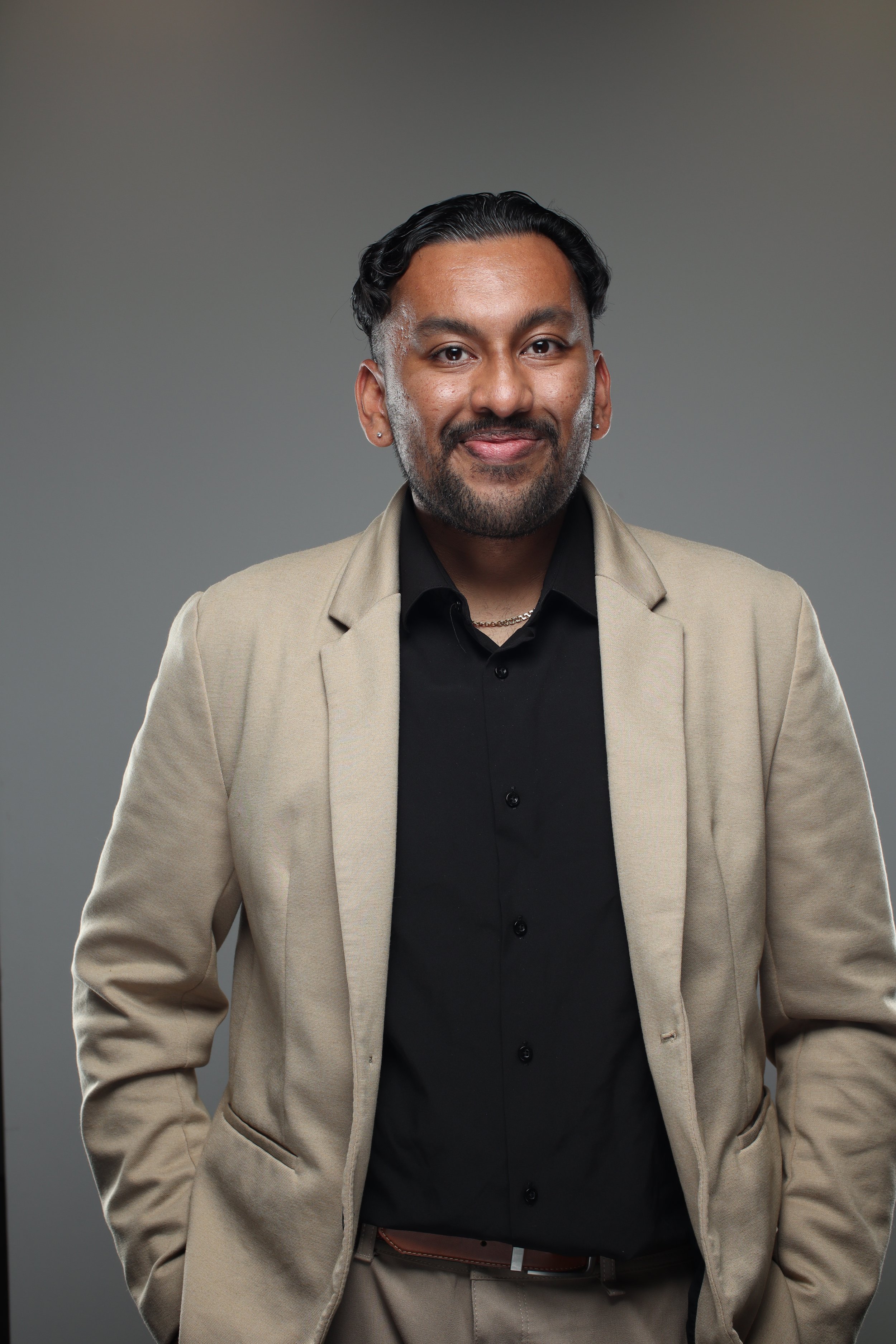 A man with a beard and mustache wearing a beige blazer, black shirt, and a gold chain, standing against a gray background.