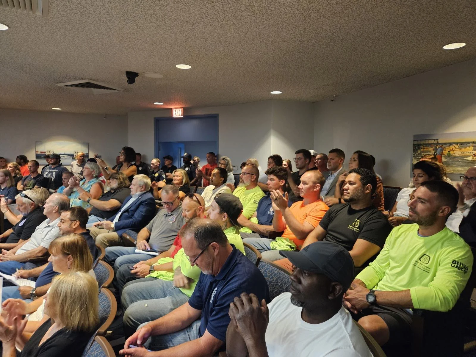 A large group of people seated indoors, attending an event or presentation; some are taking notes, others are clapping or listening attentively.
