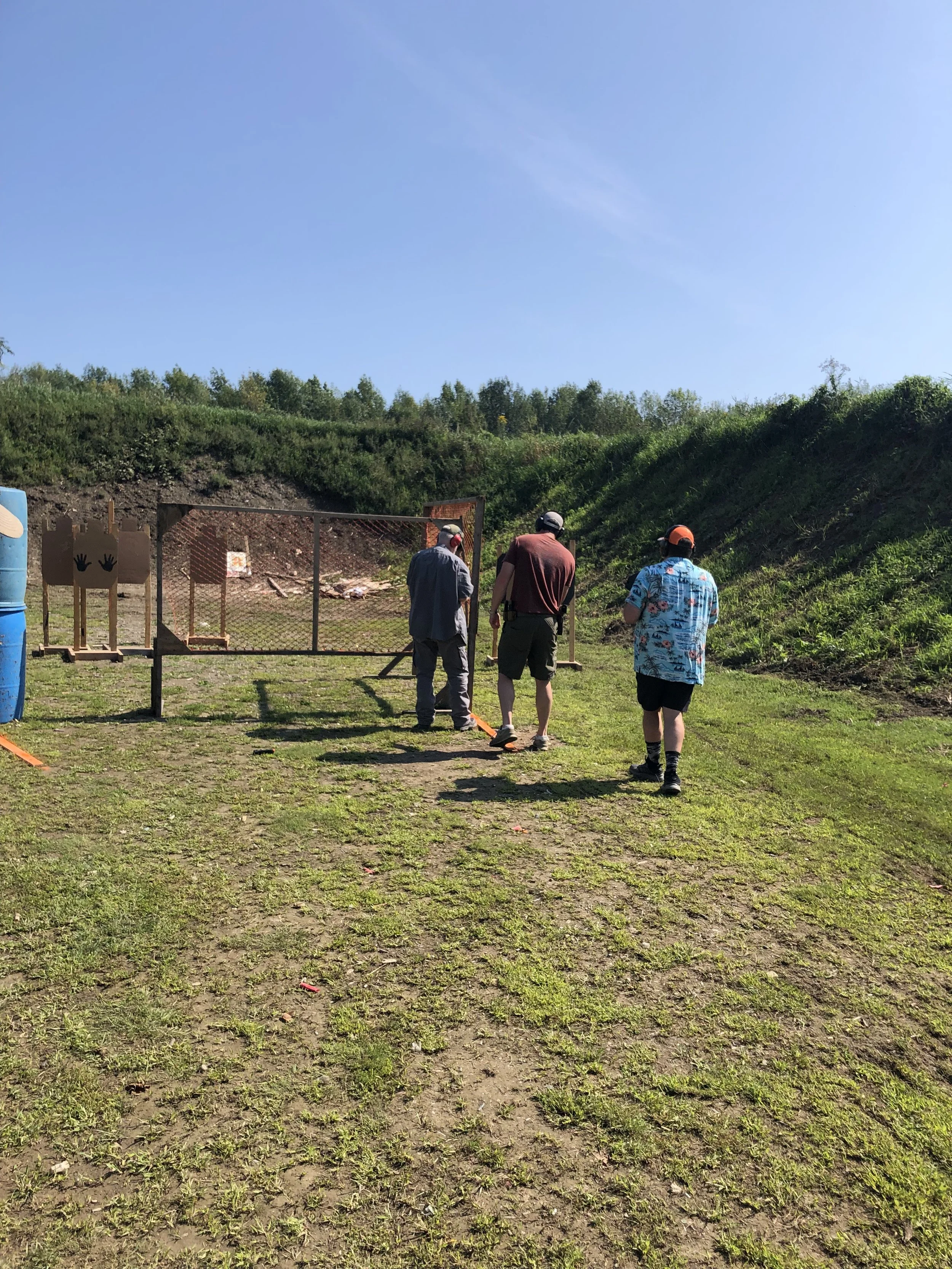 Three people at an outdoor shooting range, aiming at targets with firearms. The range has a grassy, dirt ground, a chain-link fence, and target stands with paper targets. The sky is clear and blue.