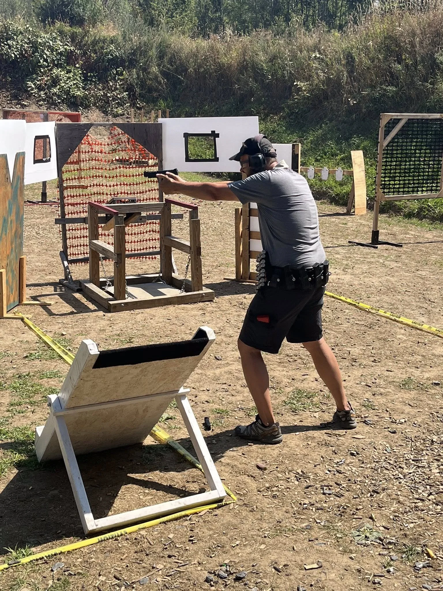 Man at outdoor shooting range aiming a handgun at targets, wearing ear protection and casual clothes.
