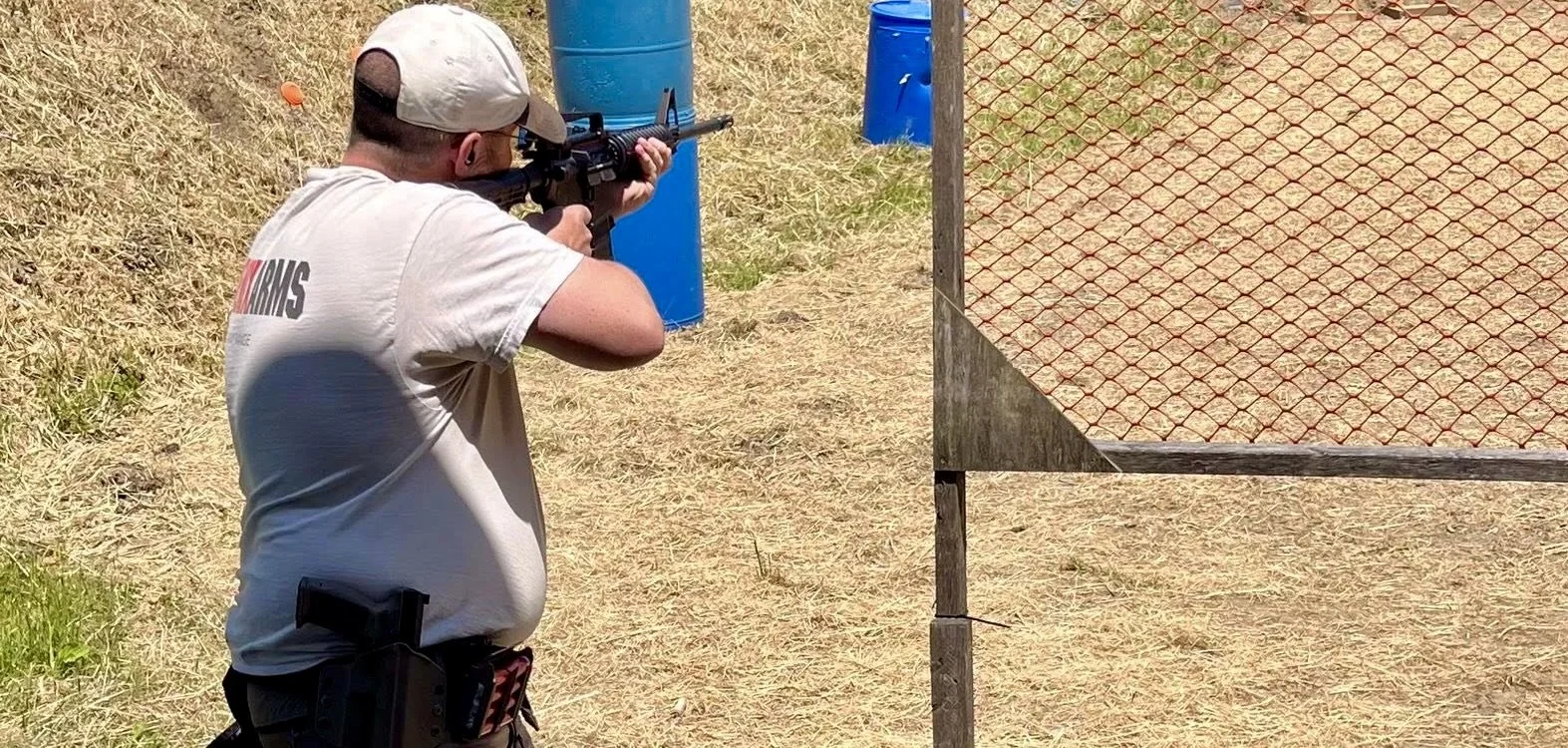 A man aiming a rifle at a shooting range, standing on dry grass, wearing a white t-shirt and a hat, with a drawer holster on his waist, next to a mesh barricade and blue barrels.