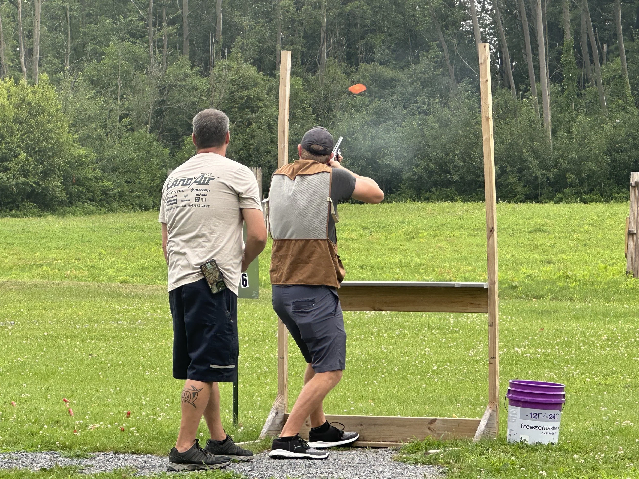 Two men participate in a shooting activity outdoors, with one aiming a gun at a target while the other observes. A purple bucket with temperature information is on the ground nearby.
