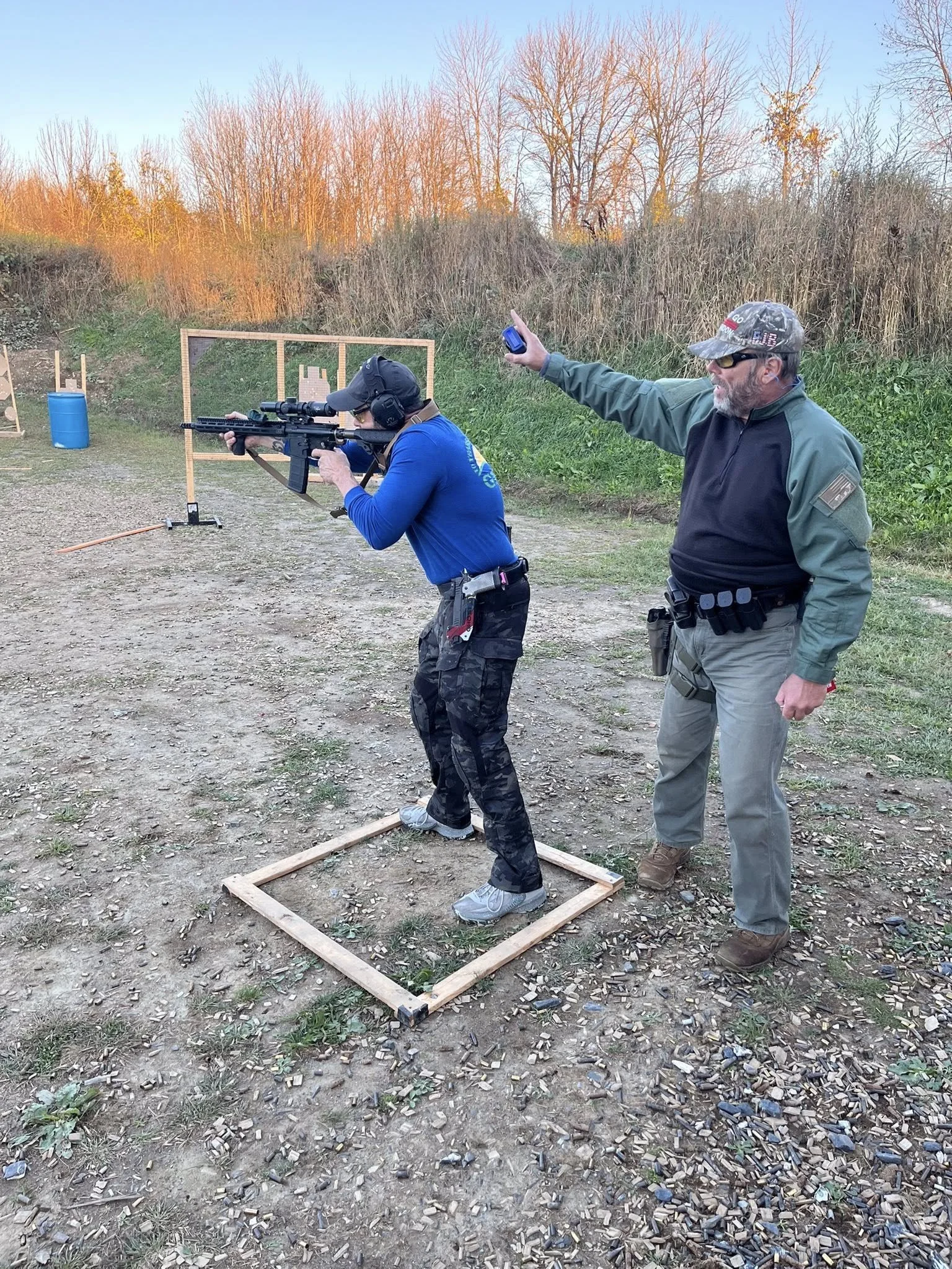 A woman in a blue shirt aiming a rifle with a scope at an outdoor shooting range, supervised by a man in a black and green jacket holding a timer, with targets and foliage in the background.