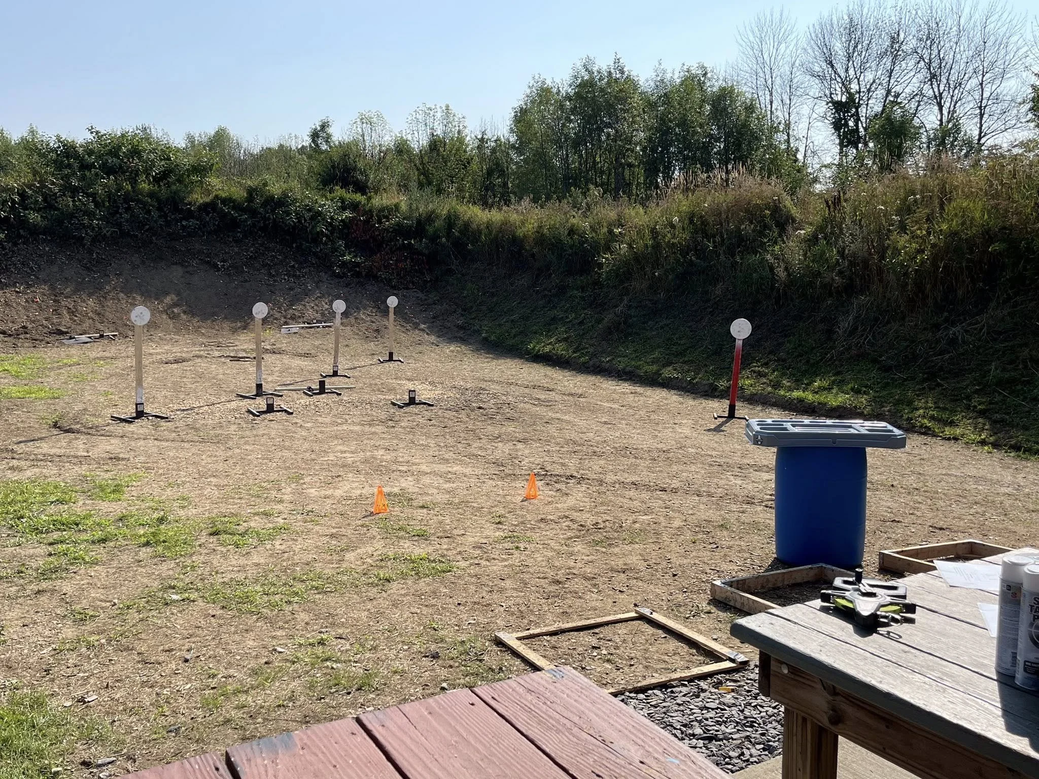 Outdoor target shooting range with paper targets on metal stands, small orange cones on ground, and drone equipment on nearby table under clear sky.