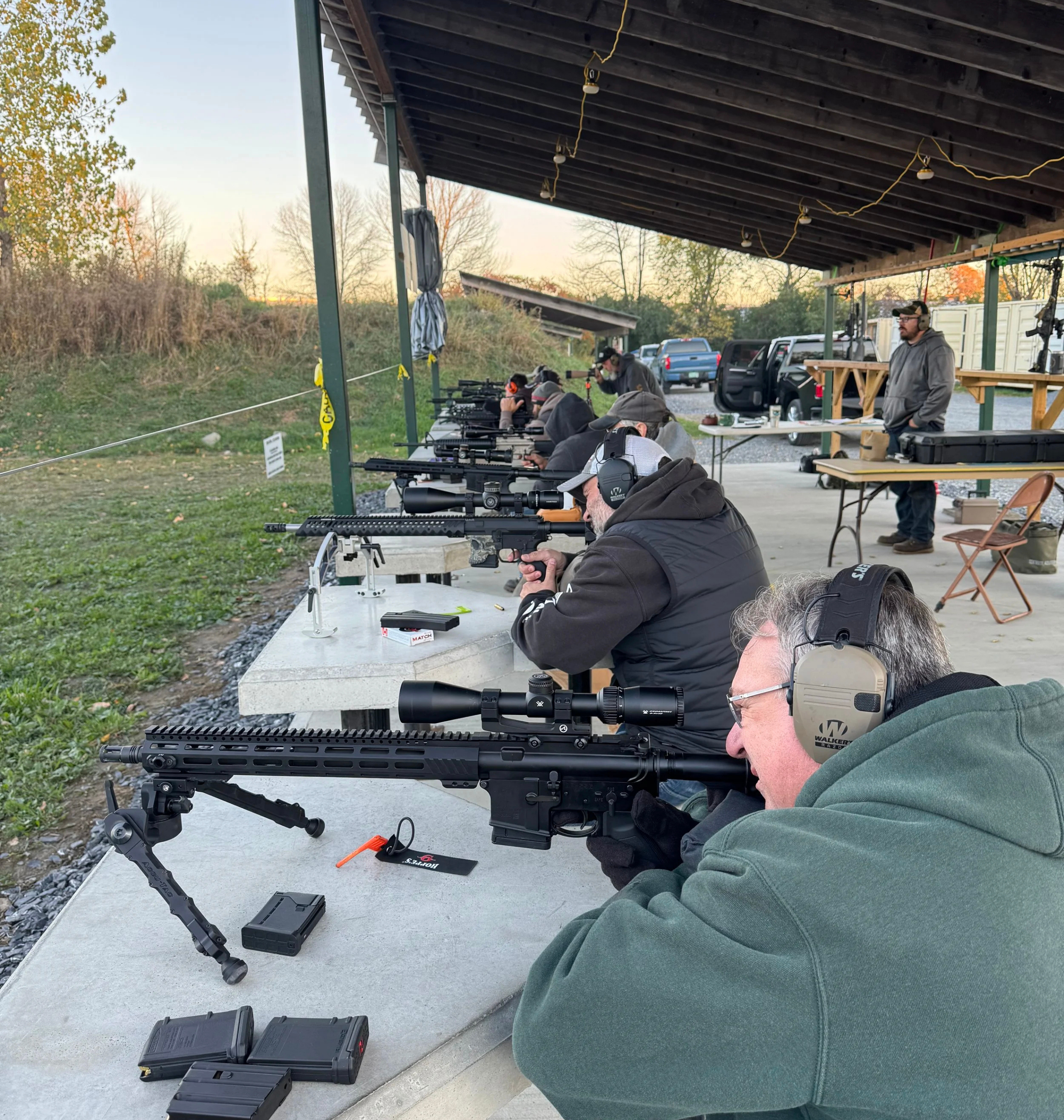 Group of people participating in a shooting session at an outdoor shooting range, aiming rifles with scopes on concrete tables under a covered structure, with some wearing headphones for hearing protection.
