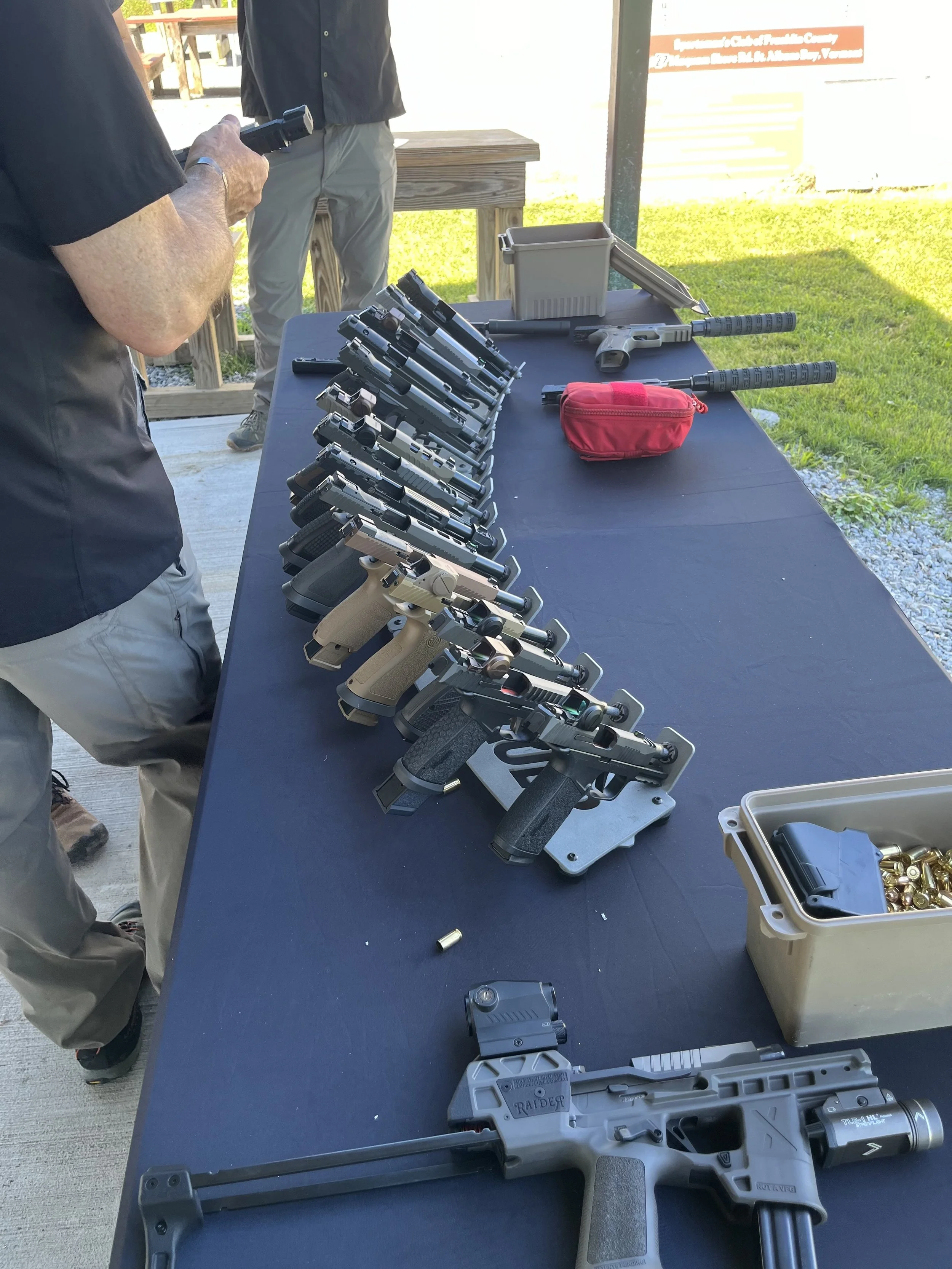 Several handguns and rifles are displayed on a black table at an outdoor shooting range, with people in the background.