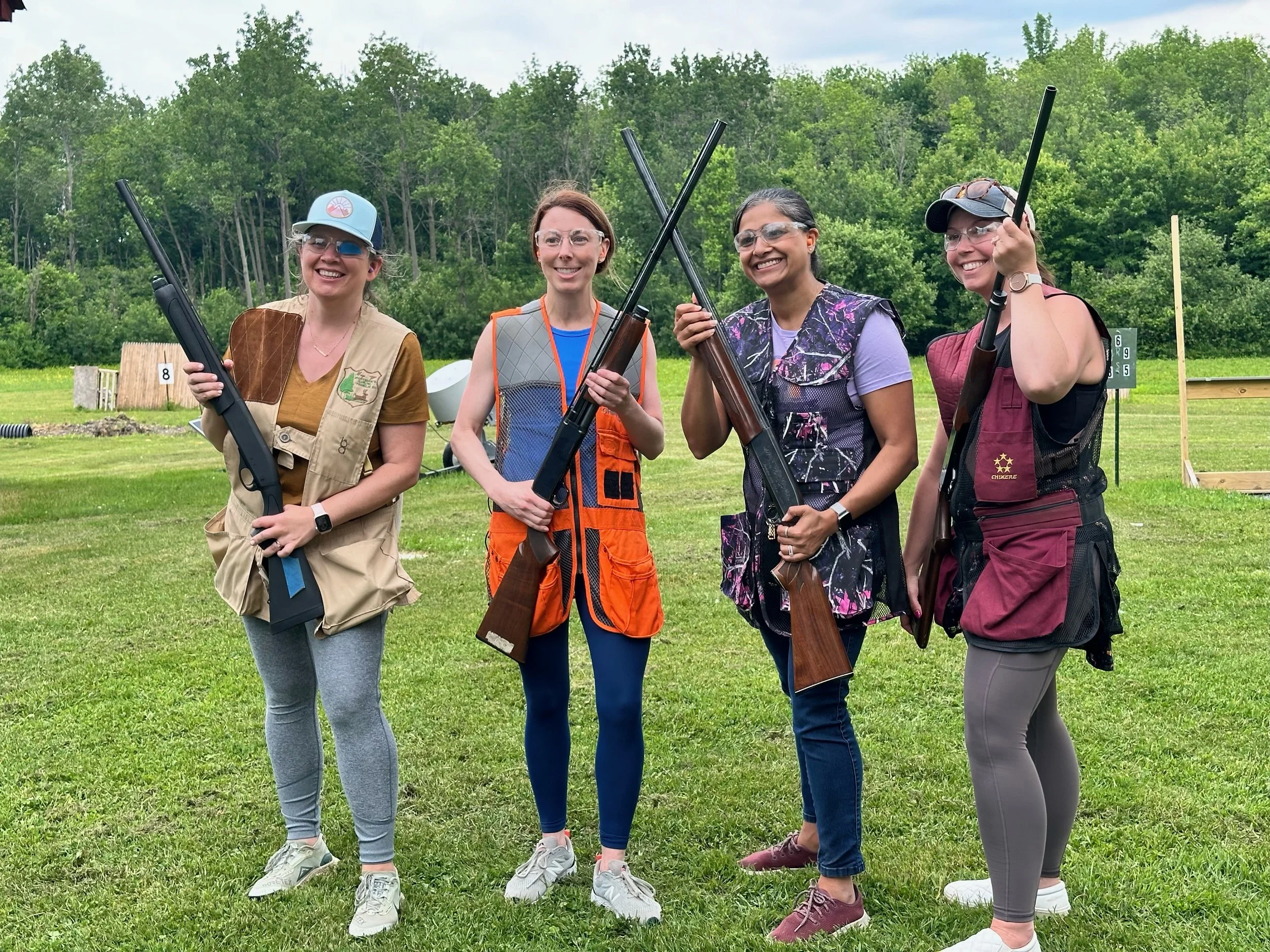 Four women holding shotguns and smiling at a shooting range surrounded by green trees.