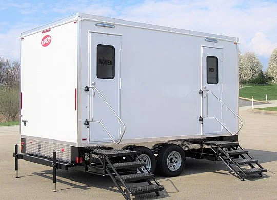White portable restroom trailer with separate doors labeled 'Women' and 'Men', equipped with stairs and handrails in an outdoor setting.