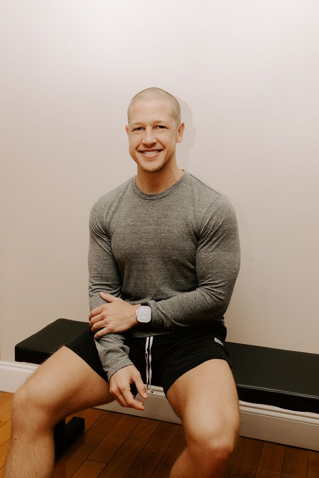 A smiling man with a shaved head, wearing a long-sleeve gray athletic shirt and black shorts, sitting on a black bench in a room with beige walls and wooden flooring.