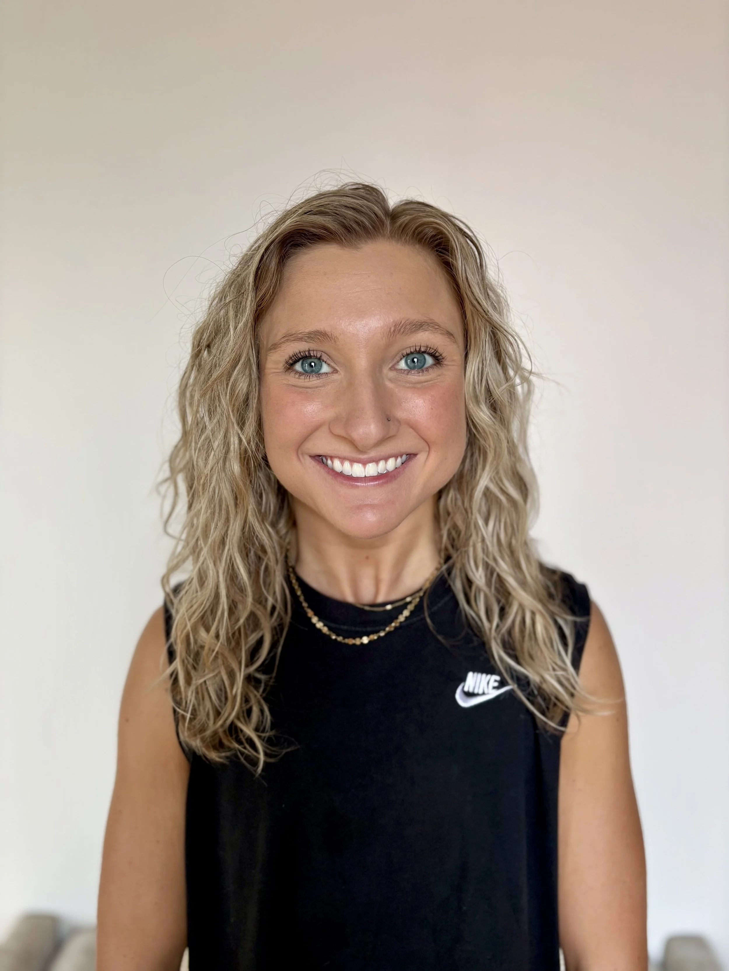 A young woman with curly blonde hair, blue eyes, and light skin, smiling in a black Nike sleeveless top wearing a layered gold necklace, standing against a plain light background.
