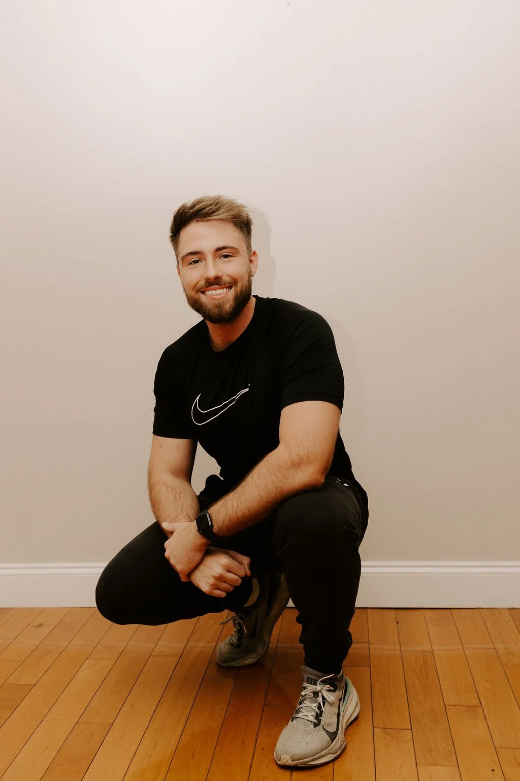 A smiling young man with a beard and short brown hair, wearing a black Nike t-shirt, black pants, and gray sneakers, crouching on one knee on a hardwood floor against a plain white wall.