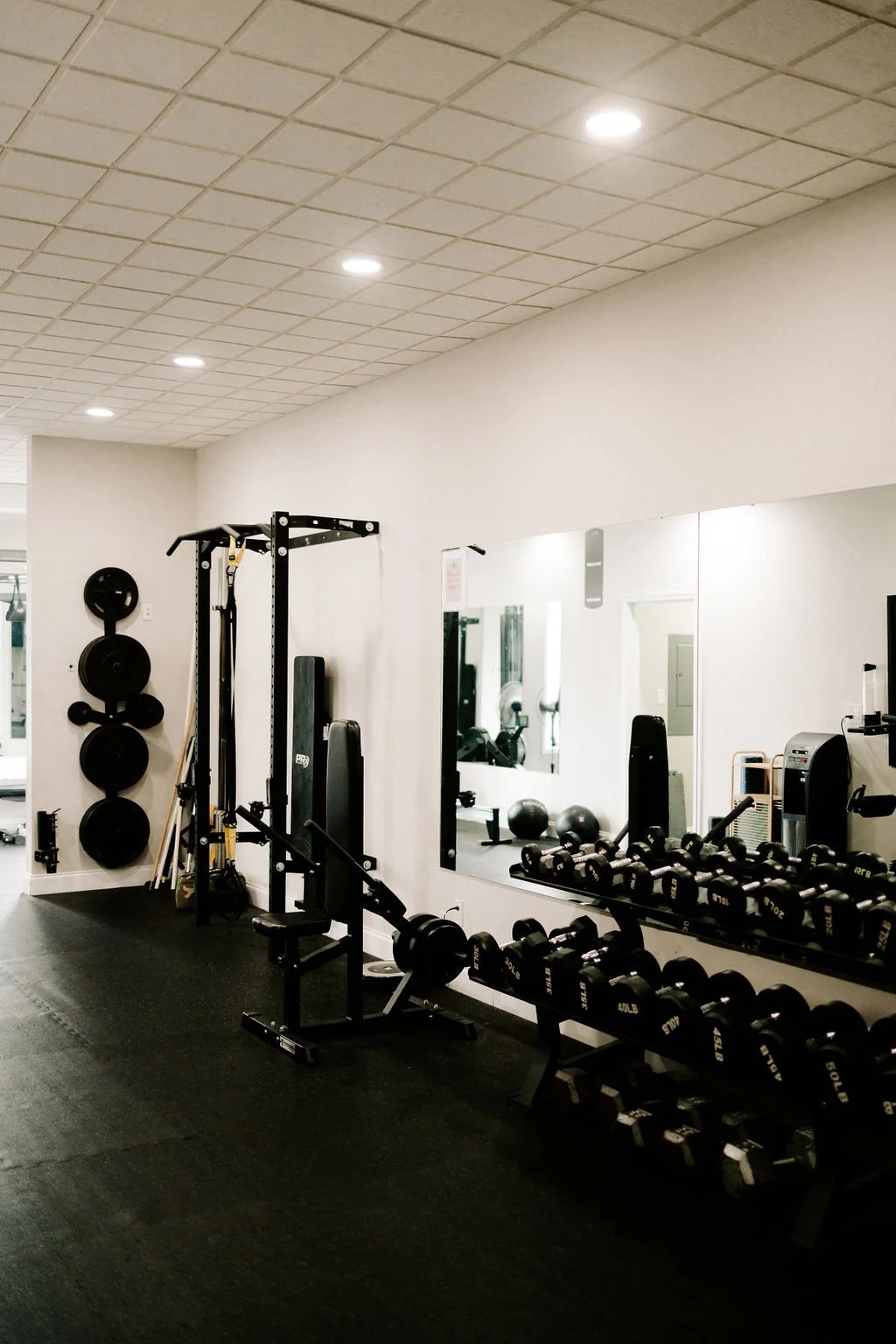 Indoor gym with weightlifting equipment including dumbbells, benches, a squat rack, medicine balls, and mirrors on the wall.