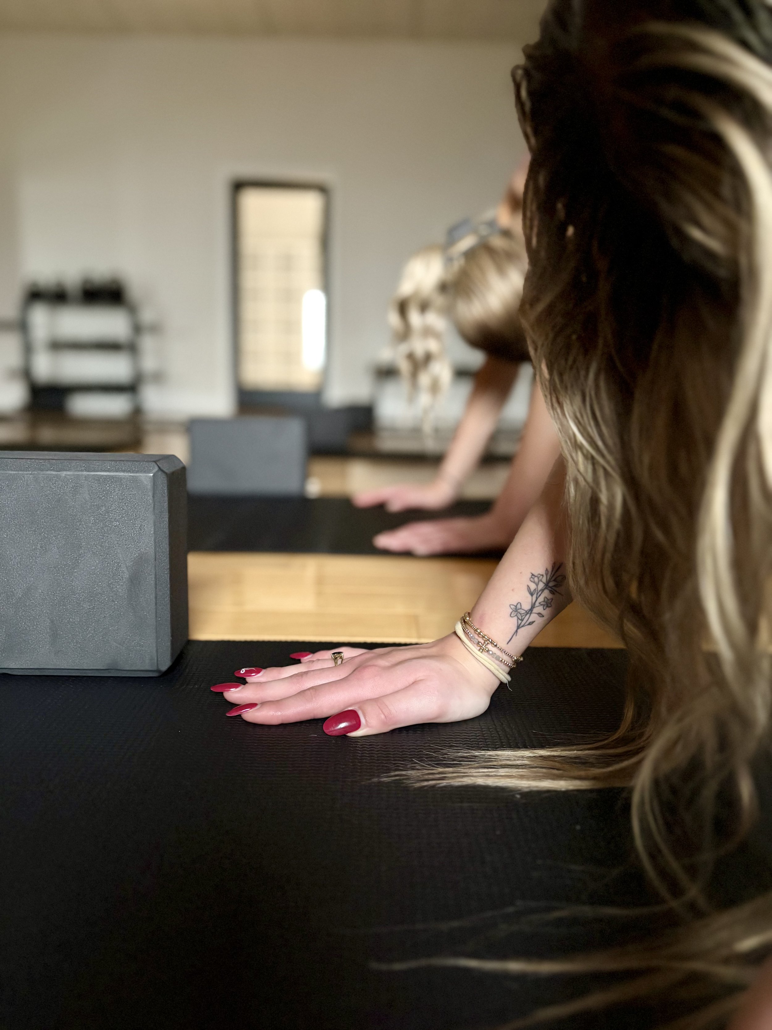 Person with red painted nails and floral tattoo on wrist practicing yoga or meditation at a black mat in a bright room with large window