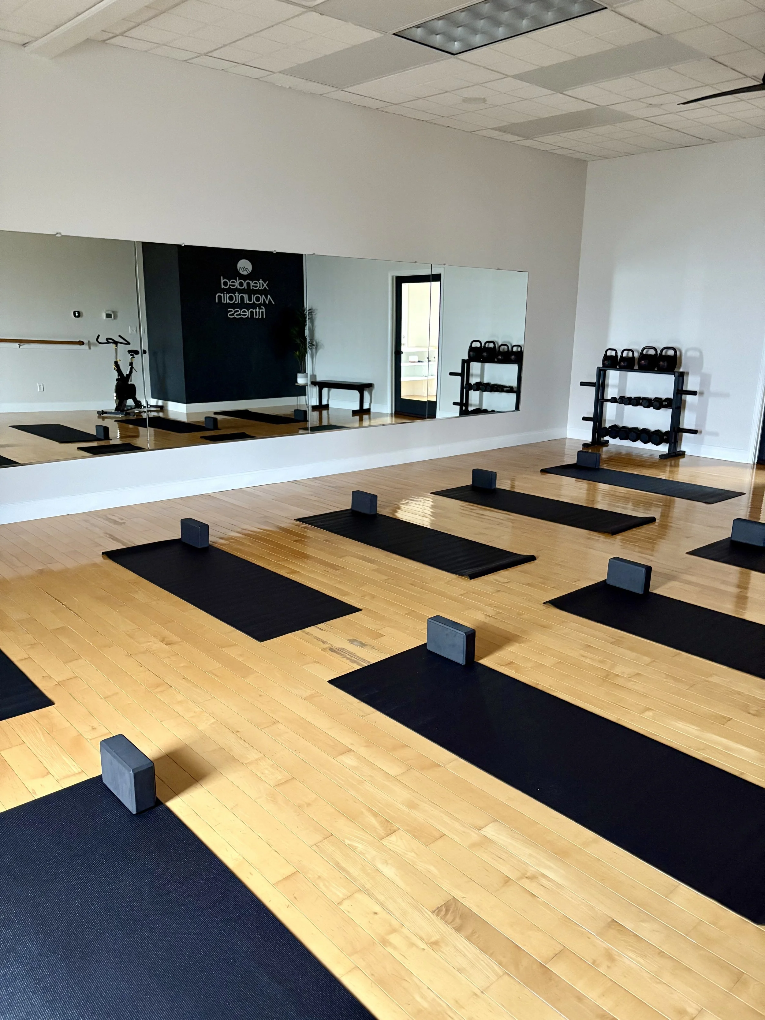 Fitness studio with black mats and yoga blocks set up for a class, mirrored wall reflecting kettlebells and a doorway to a small room.