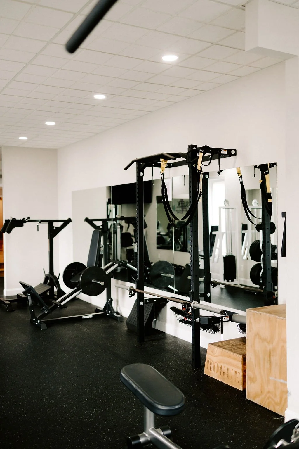 Indoor gym with black weightlifting equipment, including a squat rack with suspension straps, a rowing machine, and a mirror on the wall.