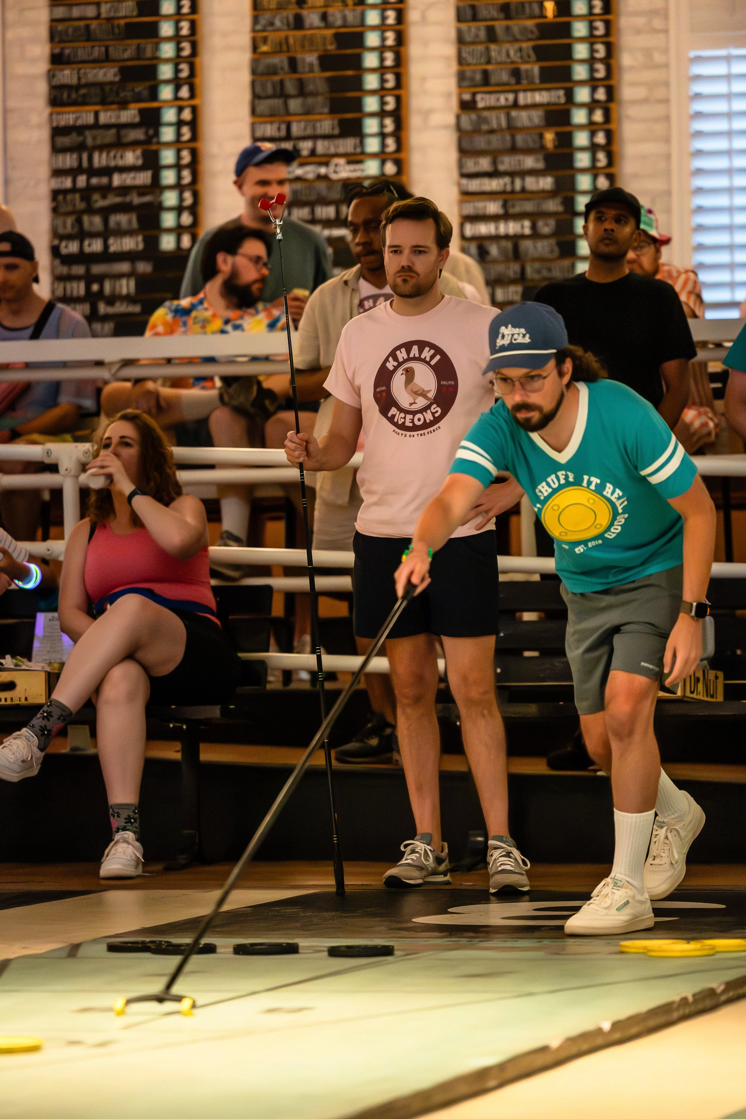 People playing shuffleboard inside a recreational area, with spectators watching from the bleachers.