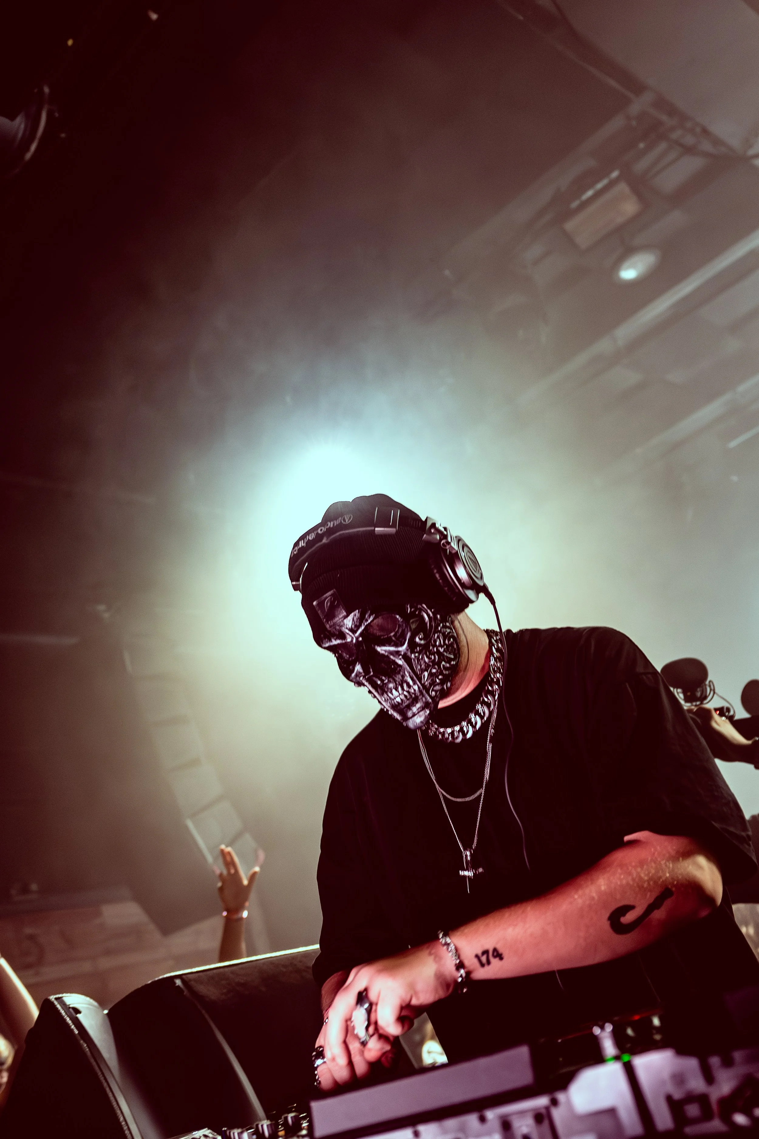 A DJ wearing a skull mask, headphones, and multiple necklaces performs at a music event, with a bright light behind them and audience members with raised hands.