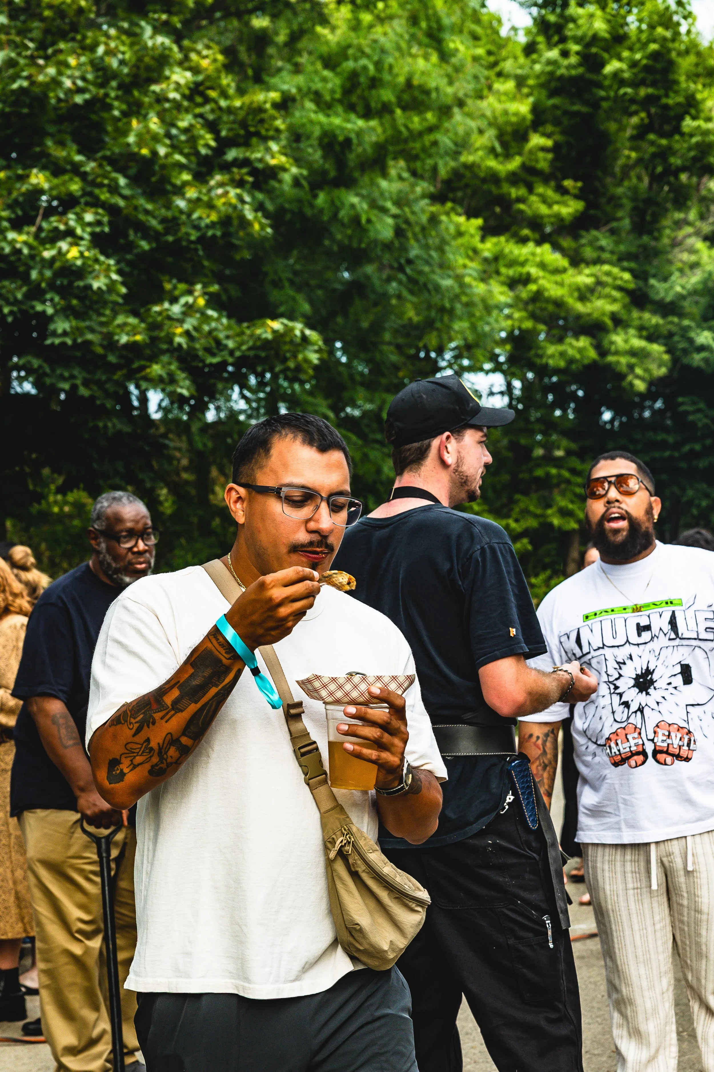 A man with tattoos and glasses eats food from a paper tray while holding a drink in an outdoor setting with trees. Other people are talking and walking nearby.