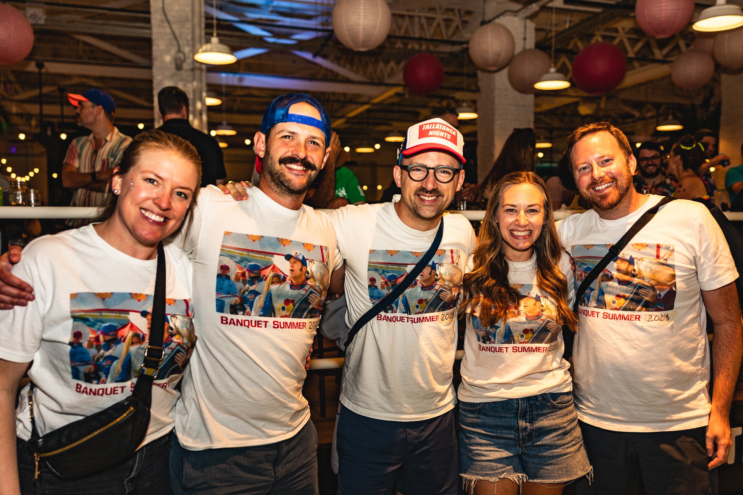Group of five smiling people wearing white T-shirts with a colorful picture and red text reading 'Banquet Summer 2025,' at a lively indoor event with hanging paper lanterns and other attendees in the background.