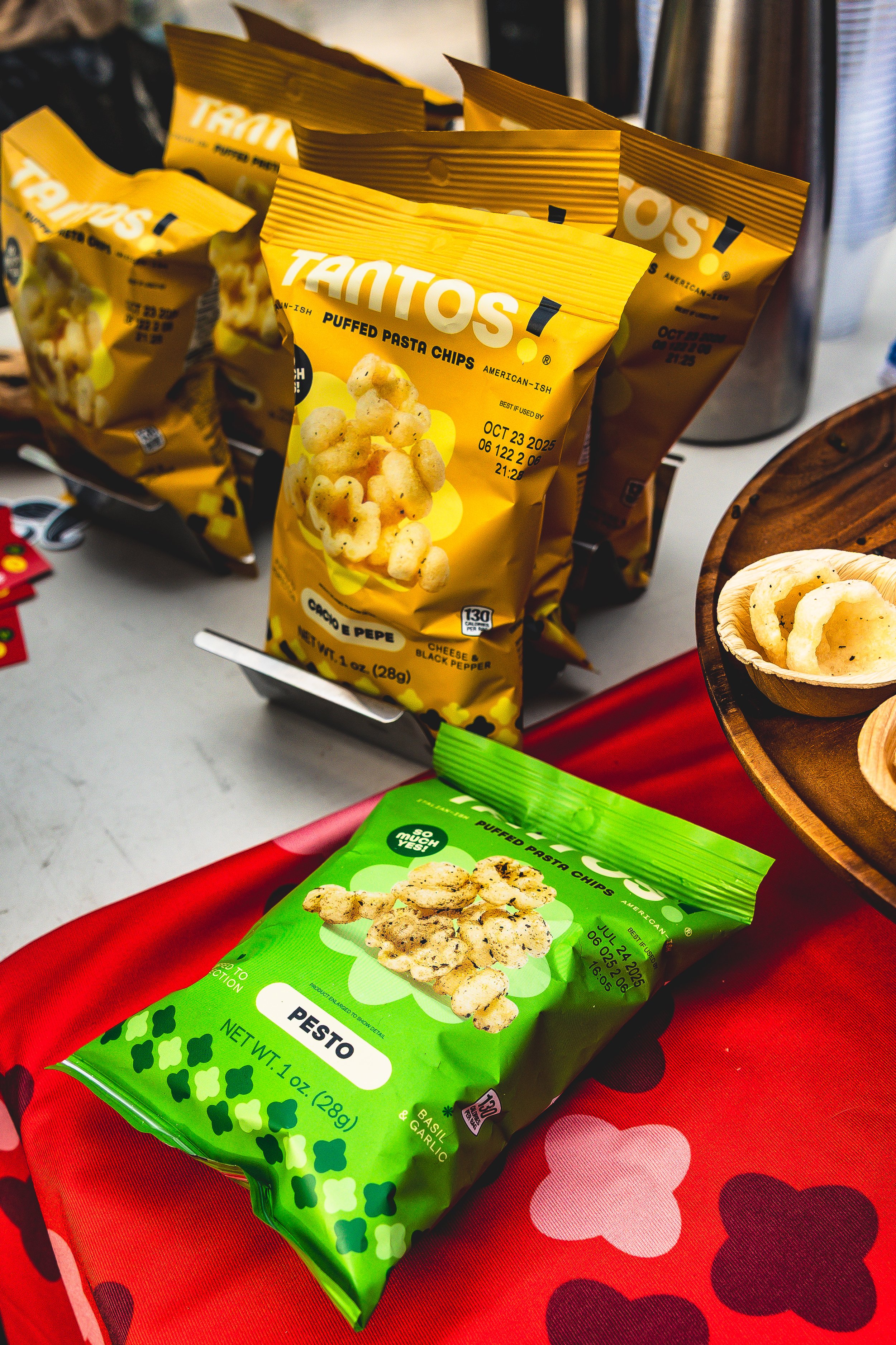 Packages of Tantos puffed pasta chips in cheese and black pepper flavor and pesto flavor on a table.