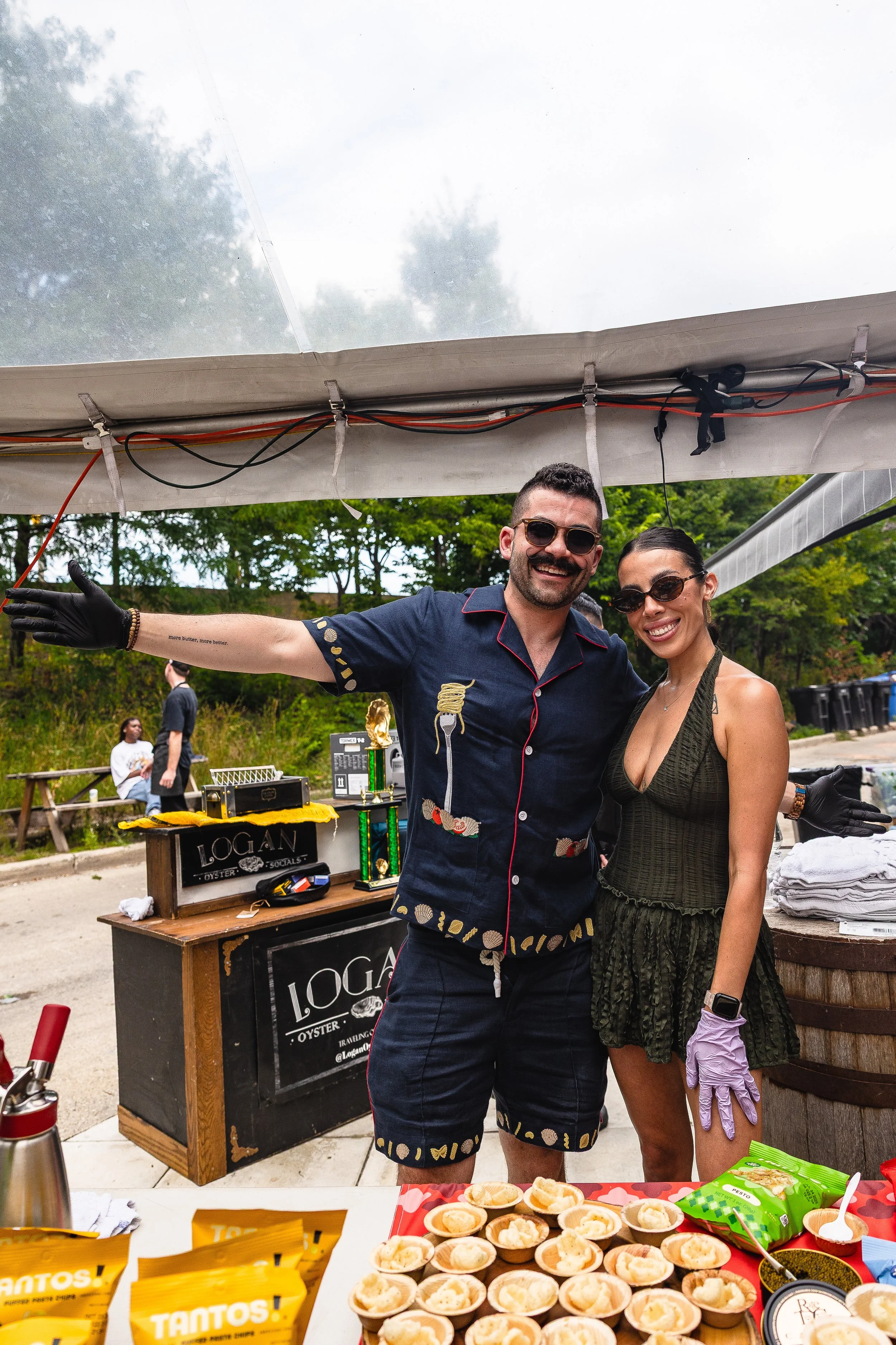 A man and woman smiling and posing at a food stand, with the man extending his arm. The food stand has trays of small bites, bags of chips, and condiments, with a sign reading 'LOGAN OYSTER SOCIAL.' The scene appears to be outdoors with trees in the 