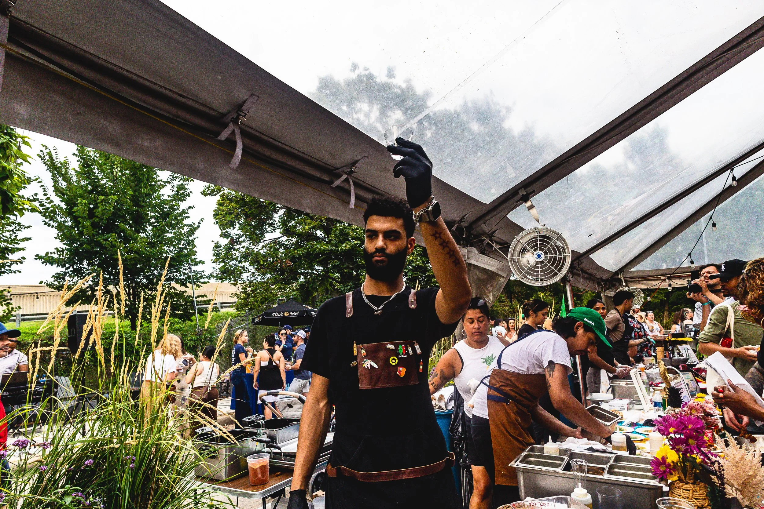 A man with a beard and tattoos, wearing a black t-shirt and black gloves, stands under a large outdoor tent at a food festival, holding a clear plastic container. Several people are visible in the background, preparing food or browsing vendors, with 