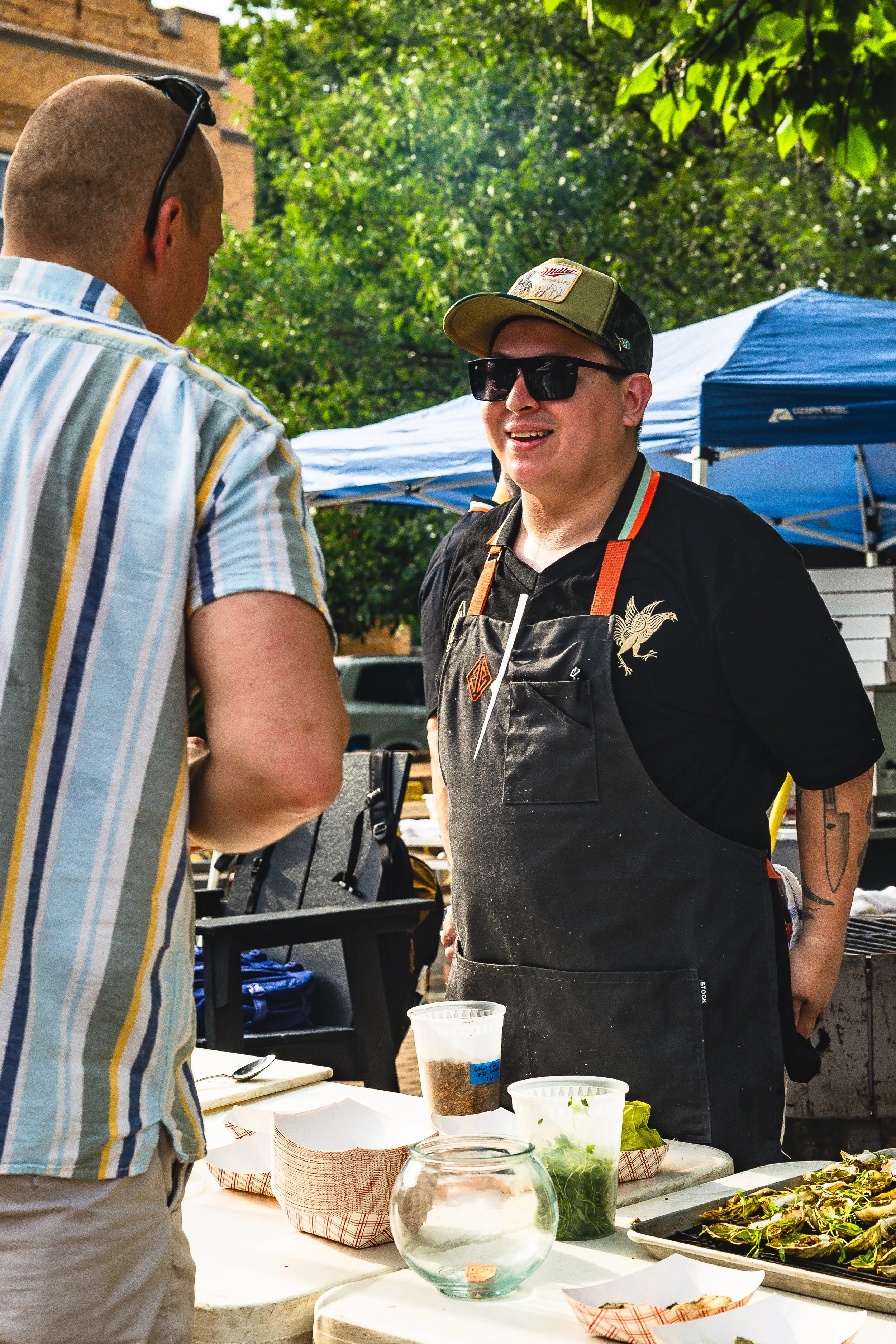 A man wearing sunglasses, a black apron with a dragon emblem, and a cap is smiling and talking to another person at an outdoor event. The table in front of him has containers with herbs, spices, and food items, and there are tents and greenery in the