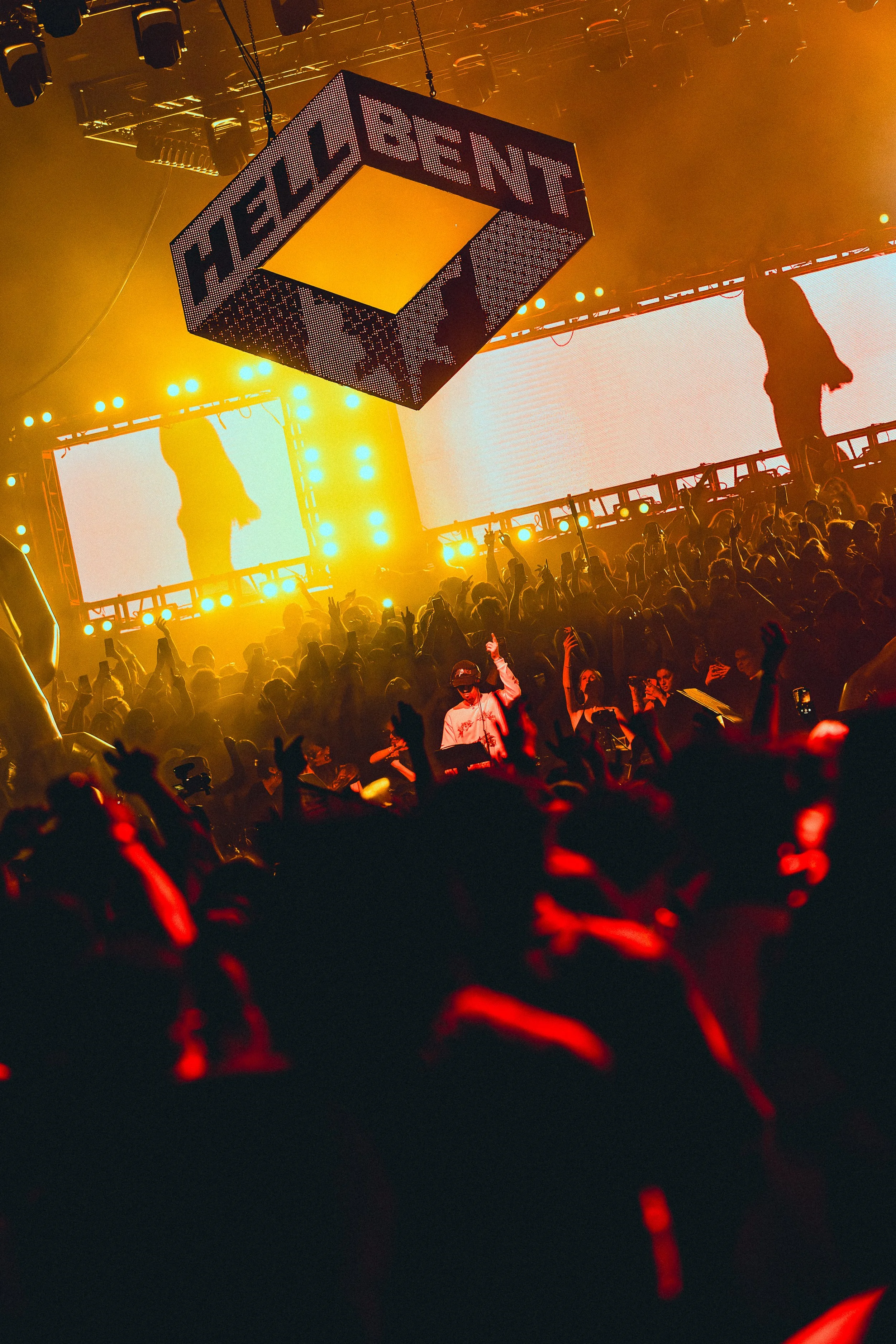 Crowd dancing at a concert with a large illuminated screen in background and yellow stage lights. There is a suspended cube-shaped sign with the text 'HELLBENT' on it.